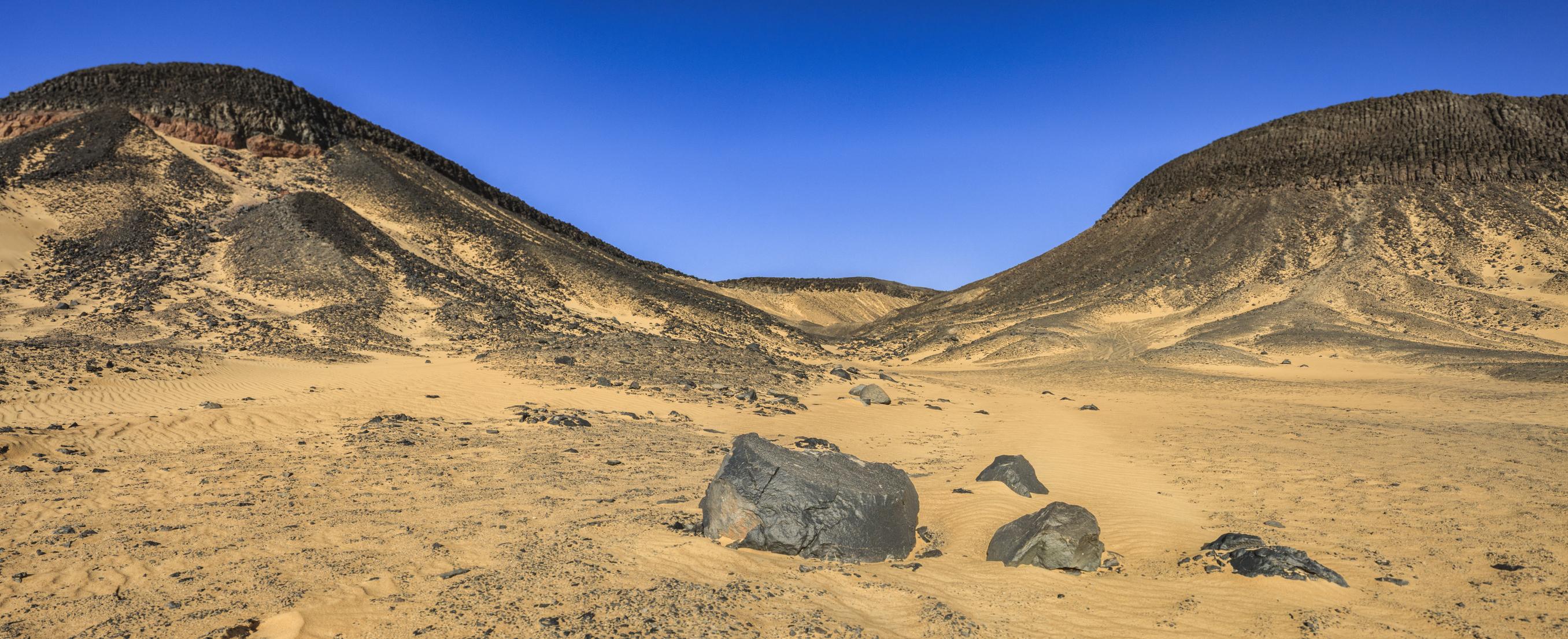 Dark volcanic rocks scattered across golden sand between two black-capped desert hills under a clear blue sky.