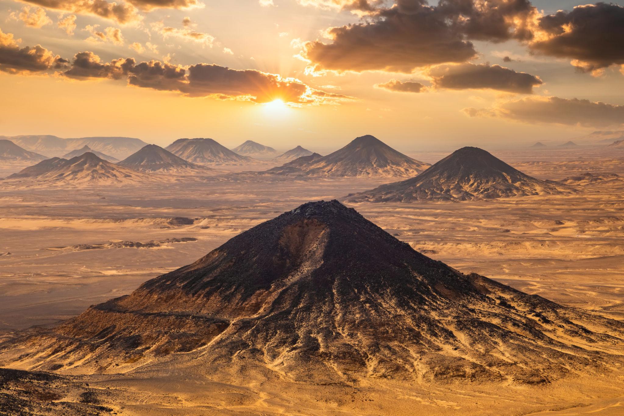 Dark volcanic cones rise from the golden Bahariya desert floor beneath a dramatic sunset sky