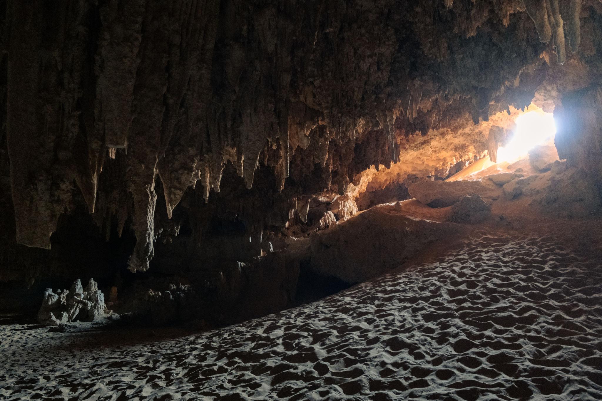 Interior of a limestone cave with stalactites on the ceiling and rippled sandy floor lit by a distant light