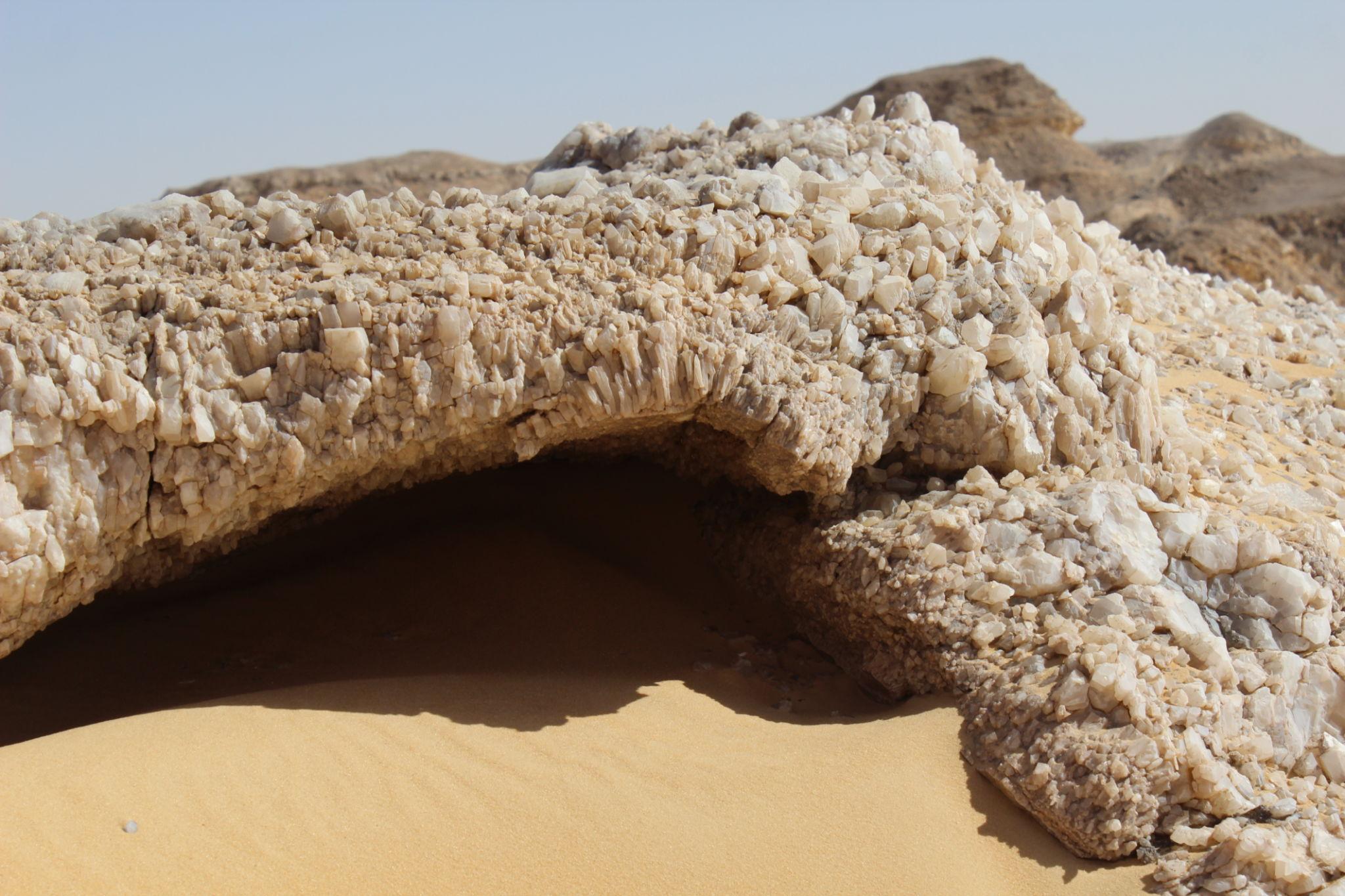 A natural arch of white crystalline rock formations rising from golden desert sand.