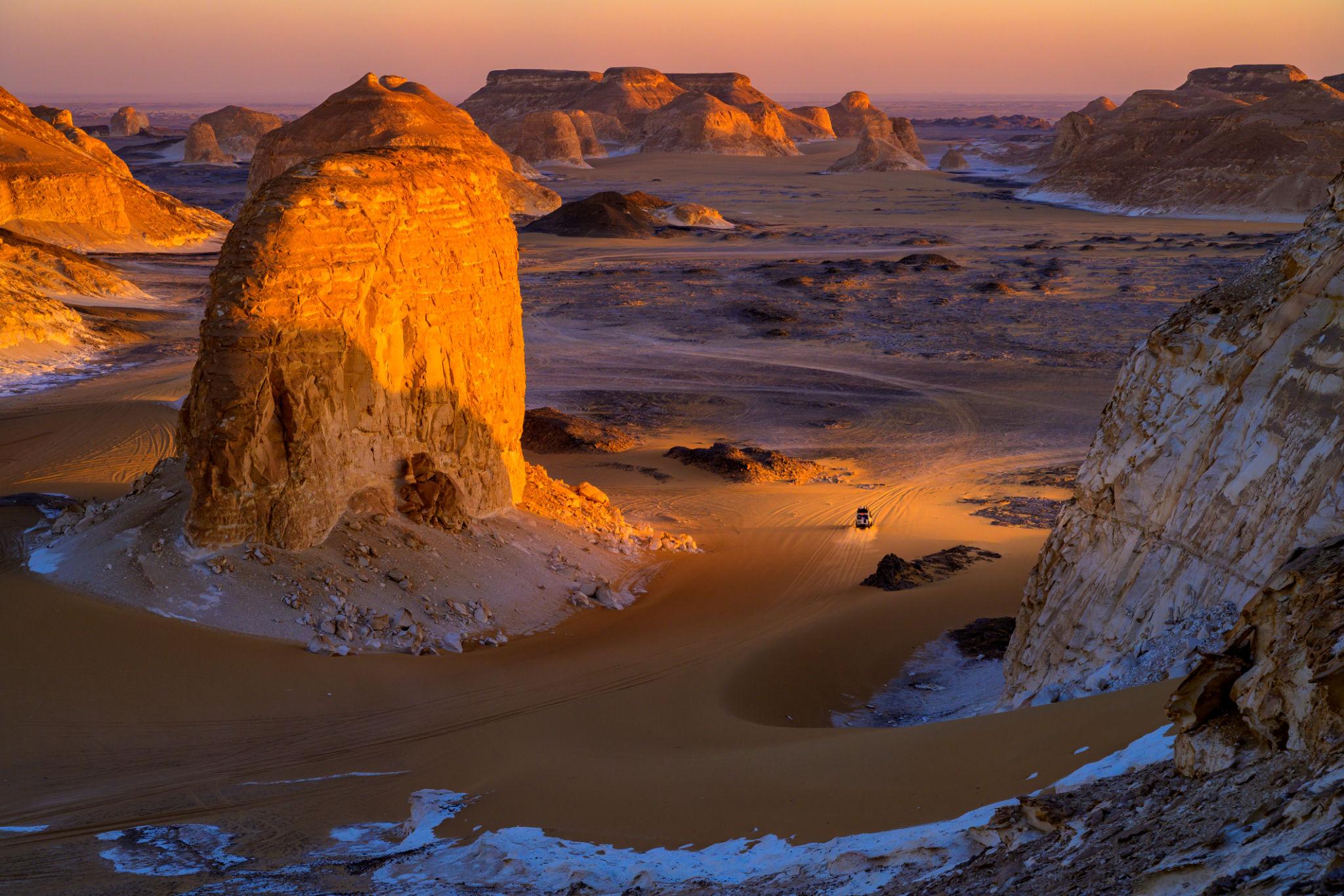 A 4x4 vehicle navigates golden sand between towering chalk rock formations at sunset