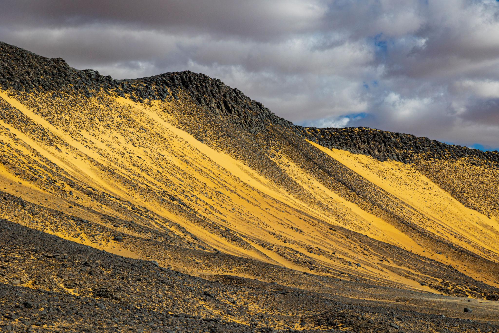 Dramatic yellow sandy streaks cutting across dark volcanic hillsides under a cloudy sky
