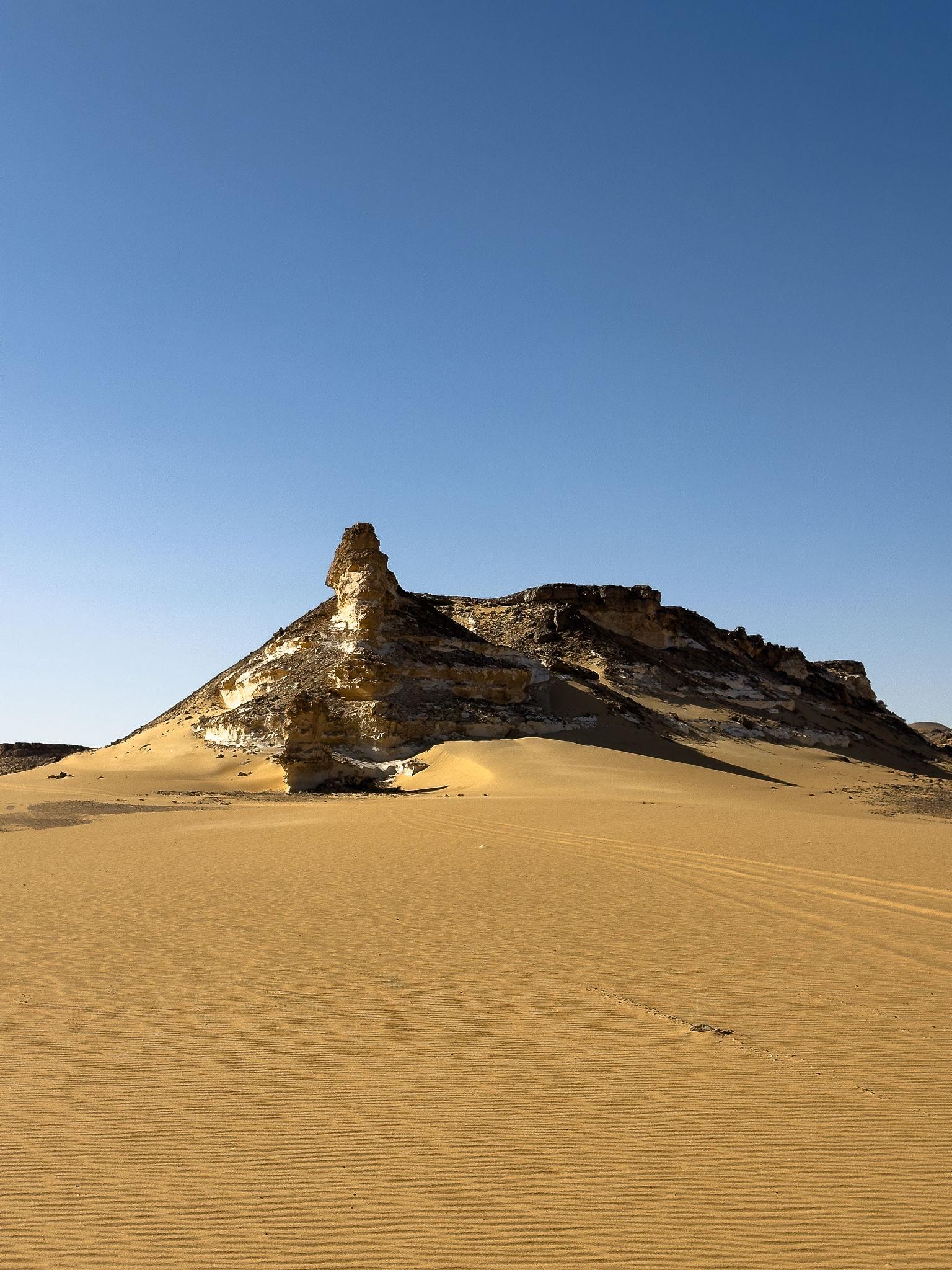 A dramatic layered rock formation rises from rippled golden sand dunes under a clear blue sky.