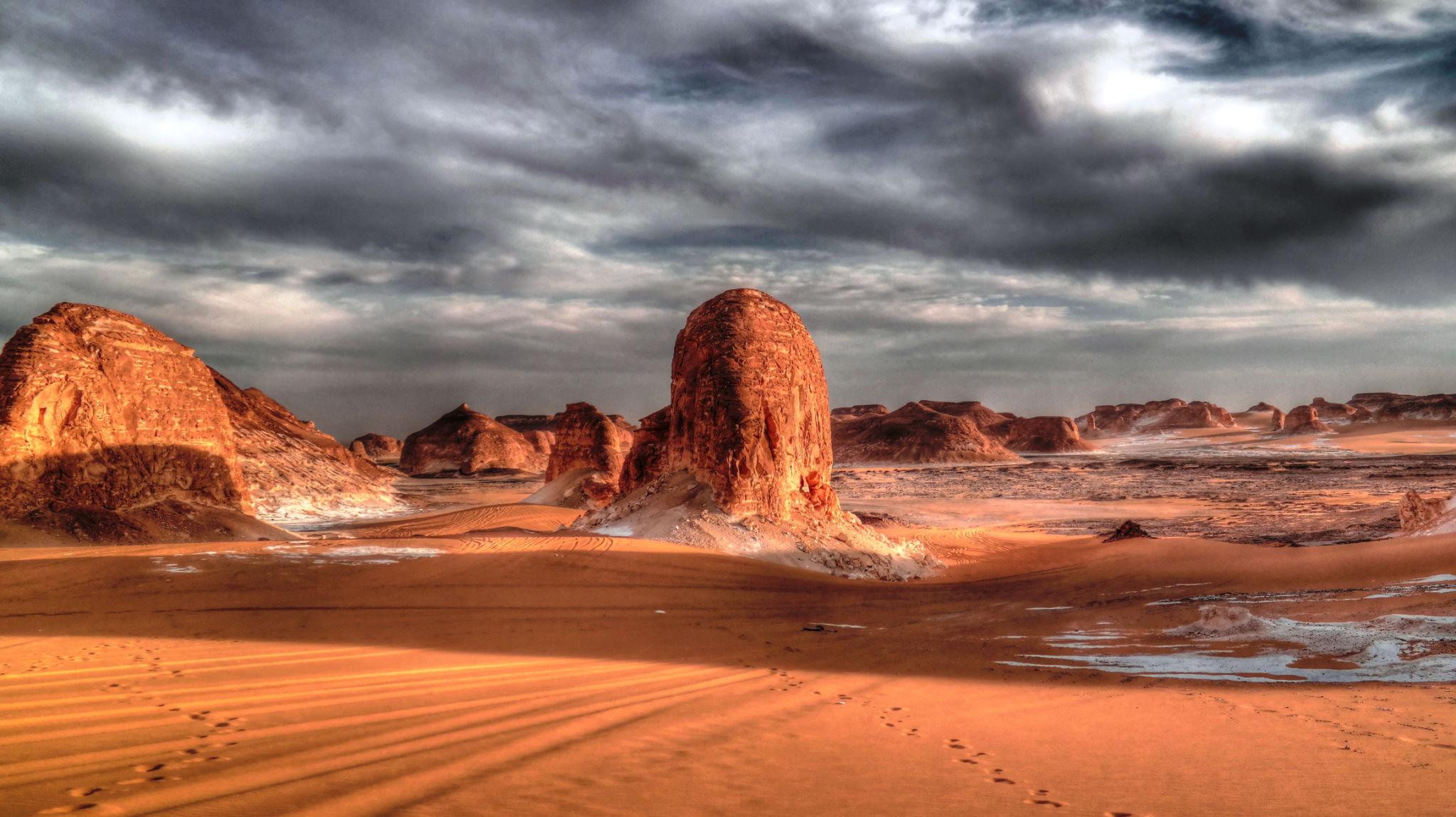 Towering red sandstone inselbergs rise from orange desert dunes under a dramatic stormy sky
