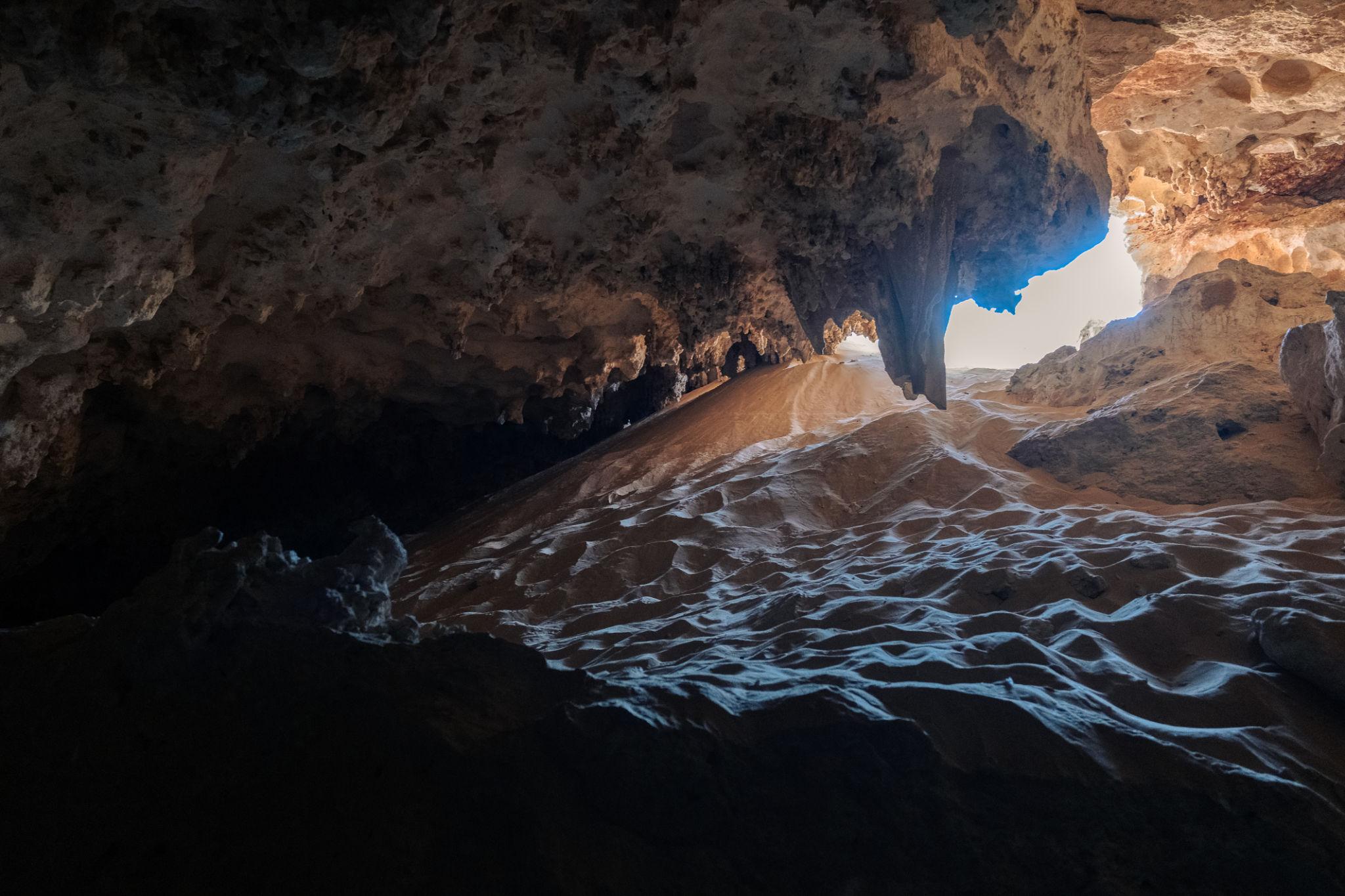 Light streams through a cave opening onto rippled sand dunes inside a limestone cavern