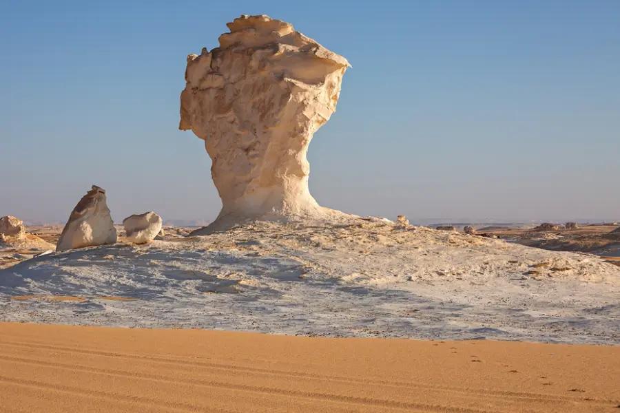 A towering white chalk inselberg rises from golden desert sands under a clear blue sky.