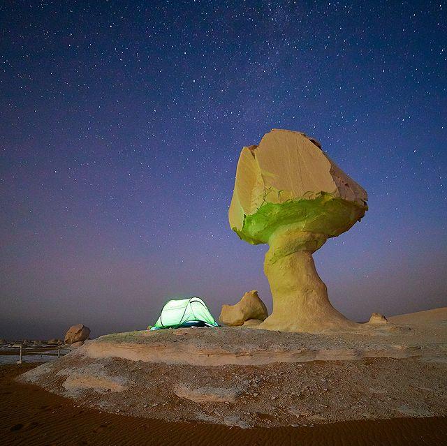 Tent glowing under a starry sky beside a chalk mushroom rock in the White Desert