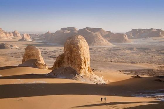 Two towering chalk rock formations rise above golden sand dunes with tiny hikers below