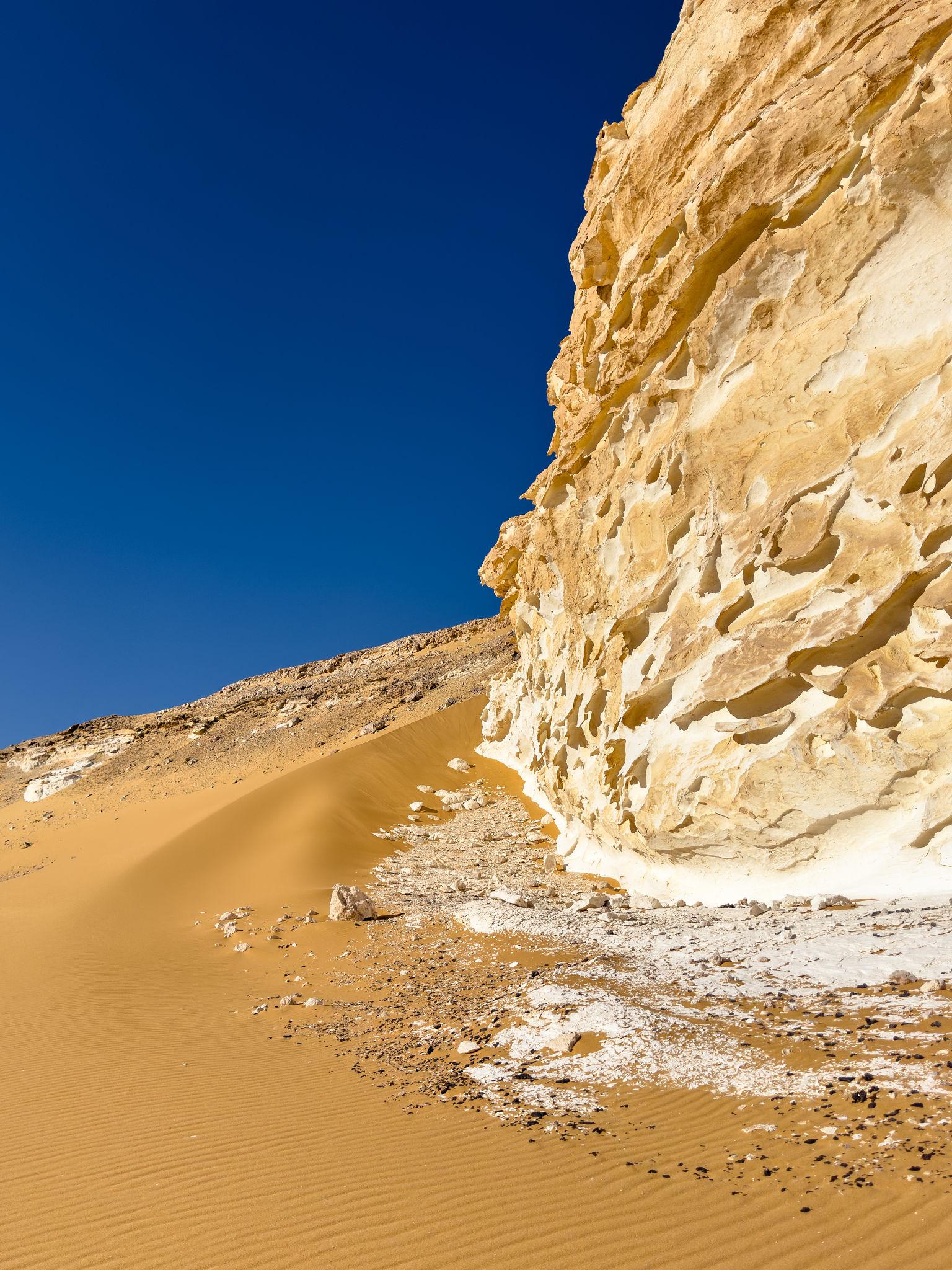 Eroded white and golden limestone cliff face rising above orange sand dunes under deep blue sky