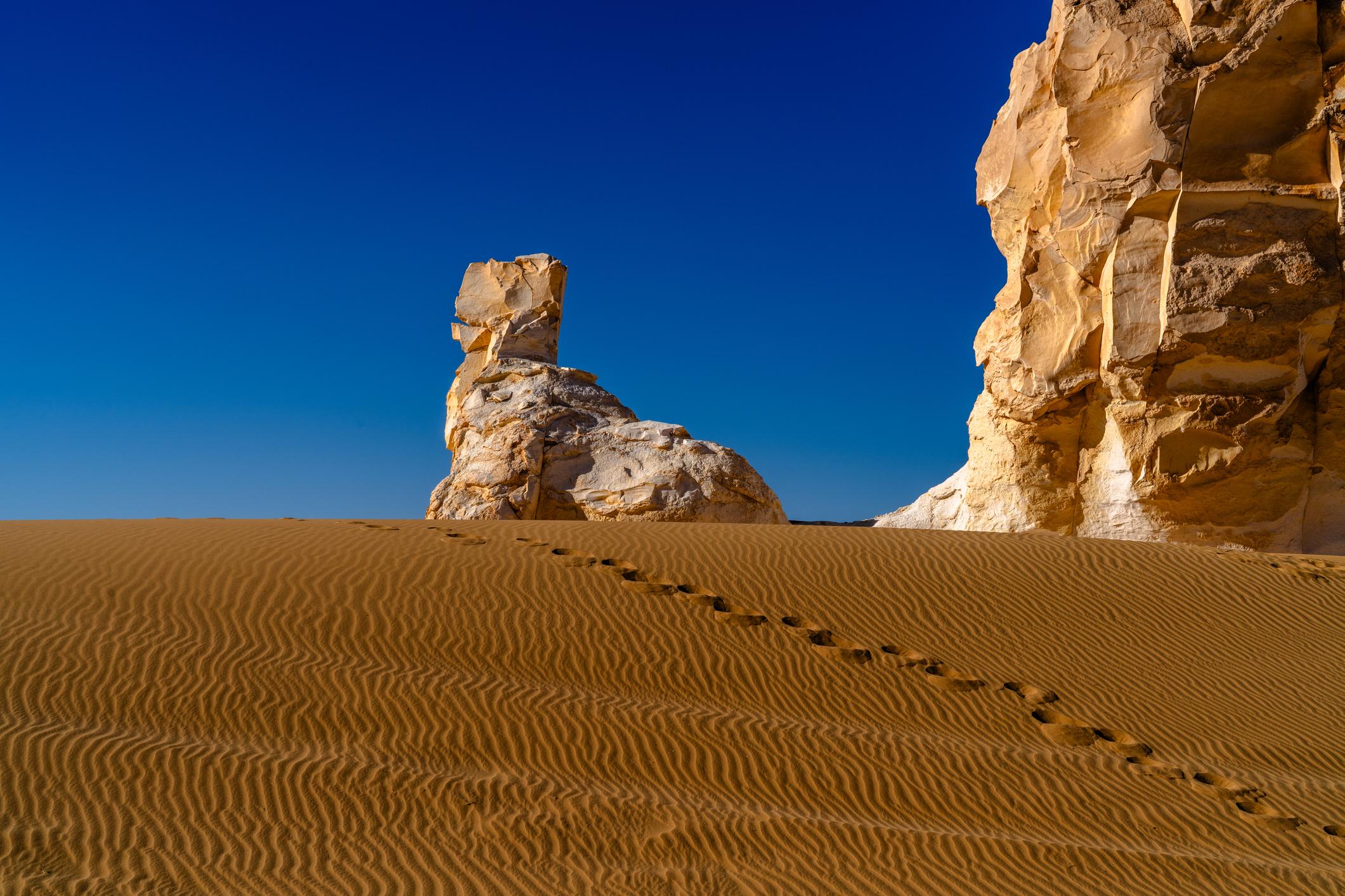 Wind-rippled sand dunes lead to eroded white limestone rock formations under a deep blue sky.