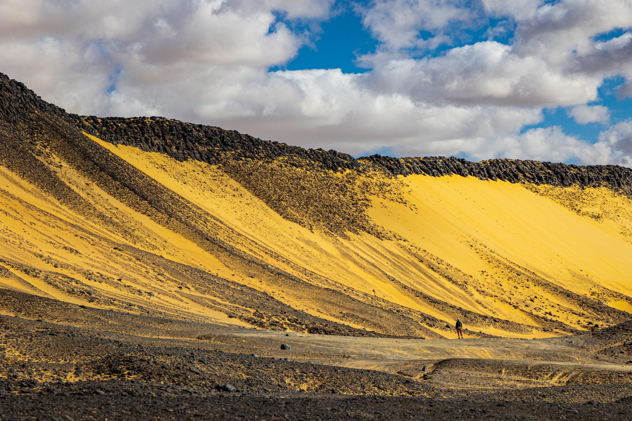 Hikers dwarfed by vast yellow sulfuric crater slopes beneath a cloudy blue sky in Bahariya Oasis