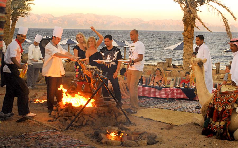Guests gather around an open beach bonfire as chefs grill meat at a Bedouin-style dinner by the Red Sea