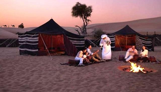 Group of tourists relaxing around a campfire beside Bedouin tents in the desert at sunset