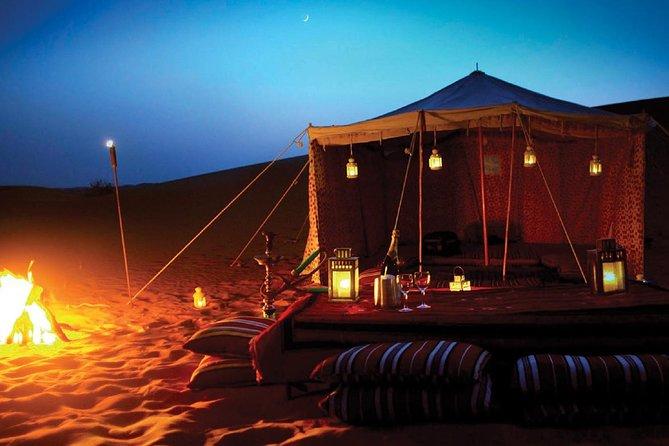 A decorated Bedouin tent glowing with lanterns beside a campfire on desert sand dunes at dusk