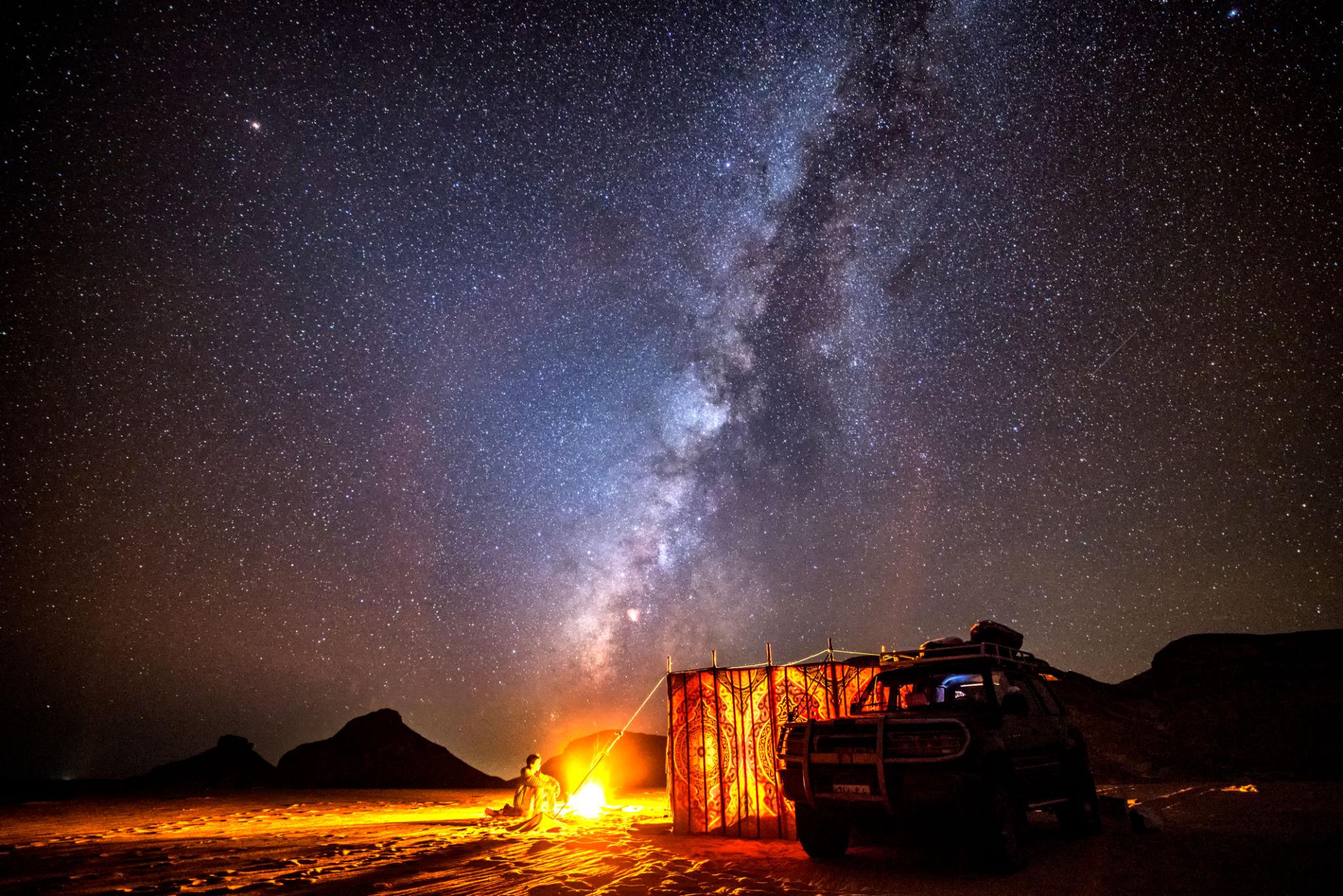 A lone camper sits by a glowing fire beside a 4x4 under a brilliant Milky Way sky in the desert