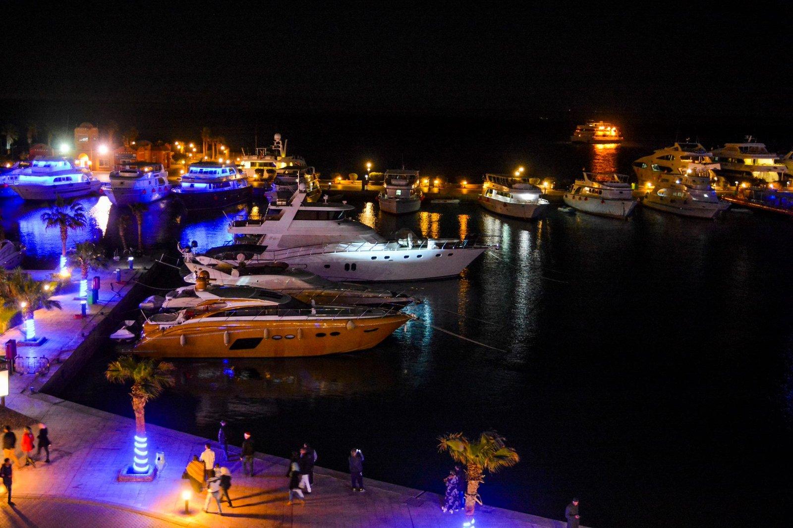 Luxury yachts moored at a glowing marina at night with people strolling the waterfront promenade