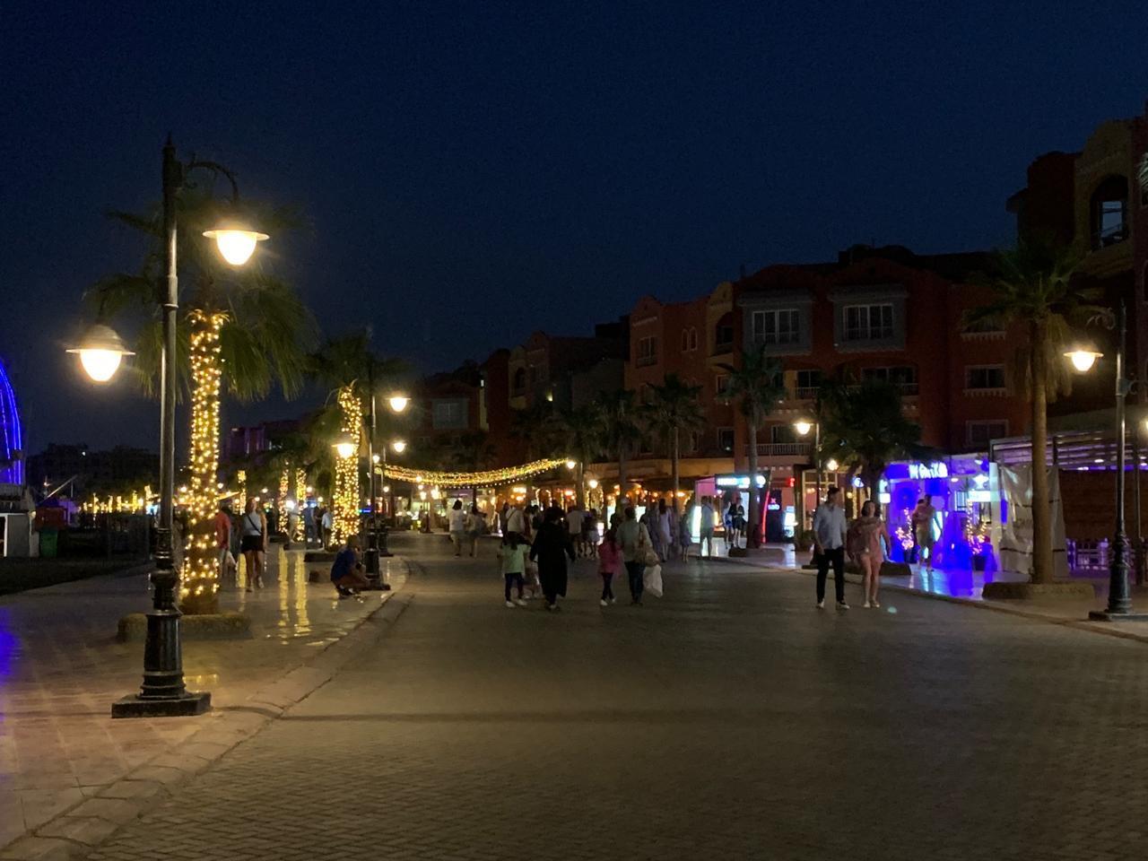 Tourists strolling along a lit waterfront promenade at night with palm trees and warm string lights