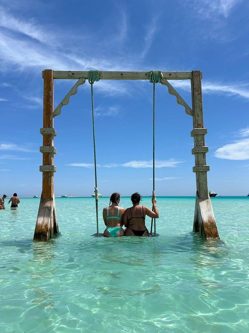 Two women sitting on a wooden swing set in the shallow turquoise waters of the Red Sea