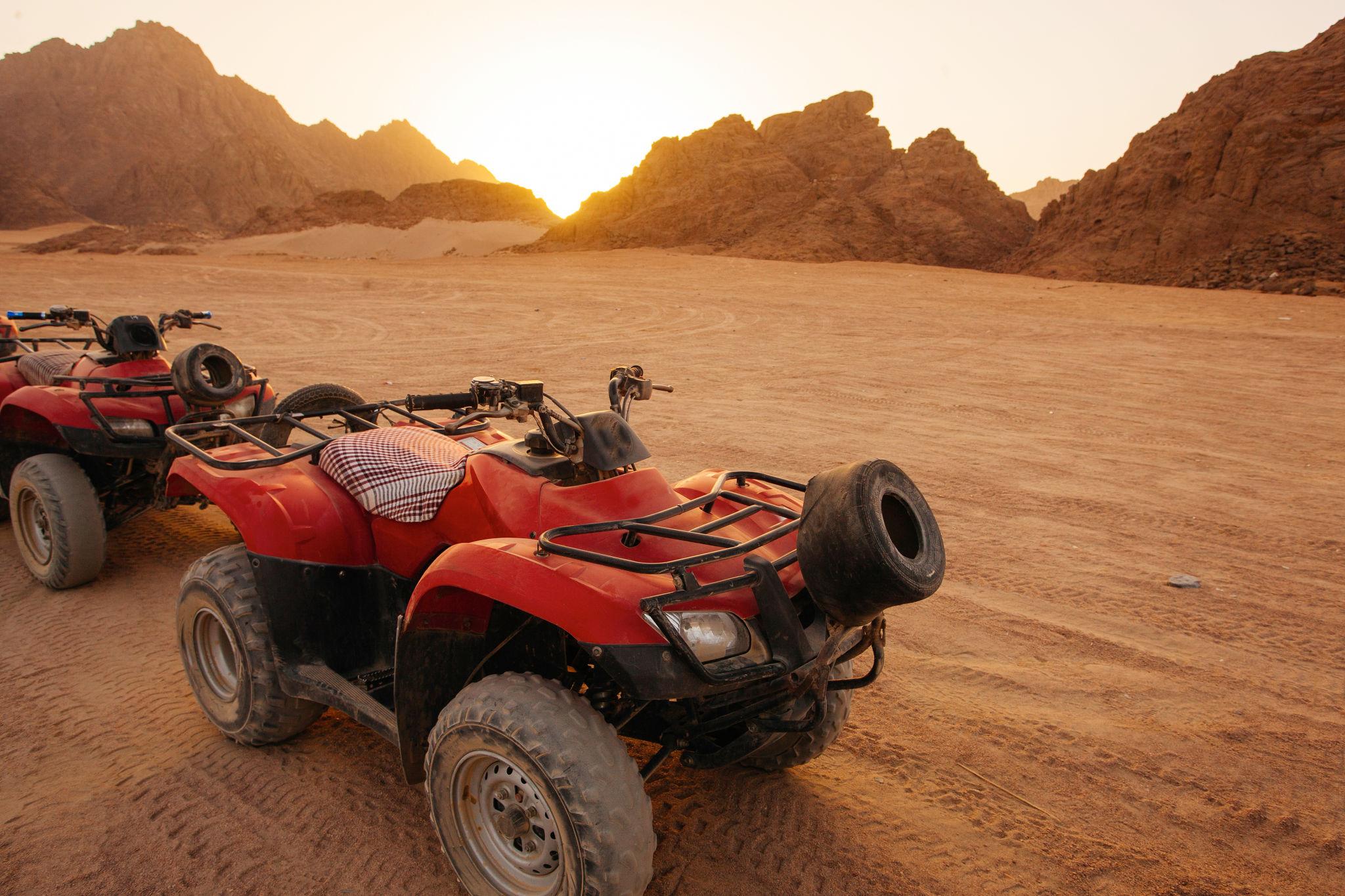 Two red quad bikes parked on sandy desert with rocky mountains at sunset