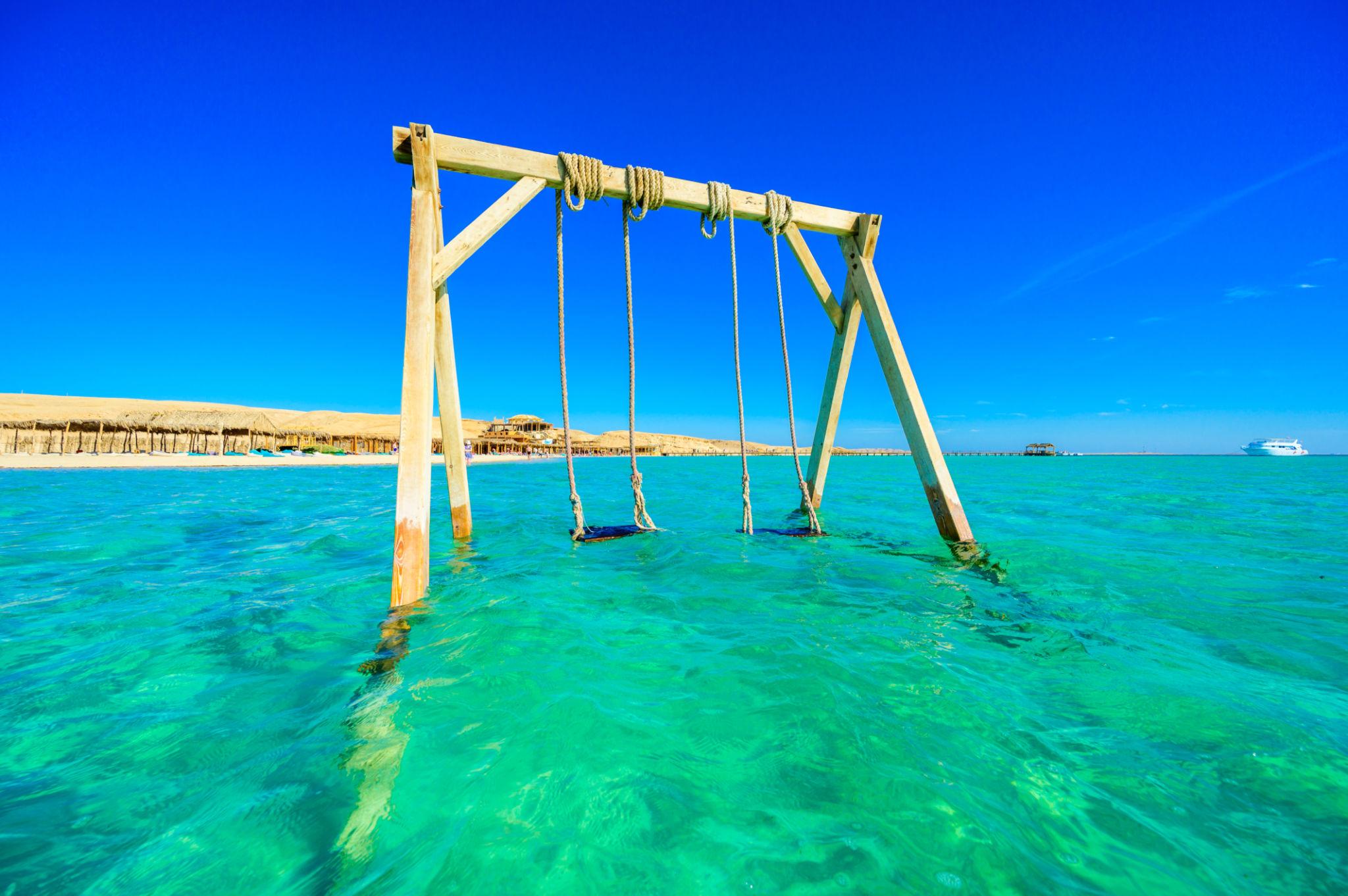 Wooden rope swings standing in clear turquoise Red Sea water with a sandy island behind
