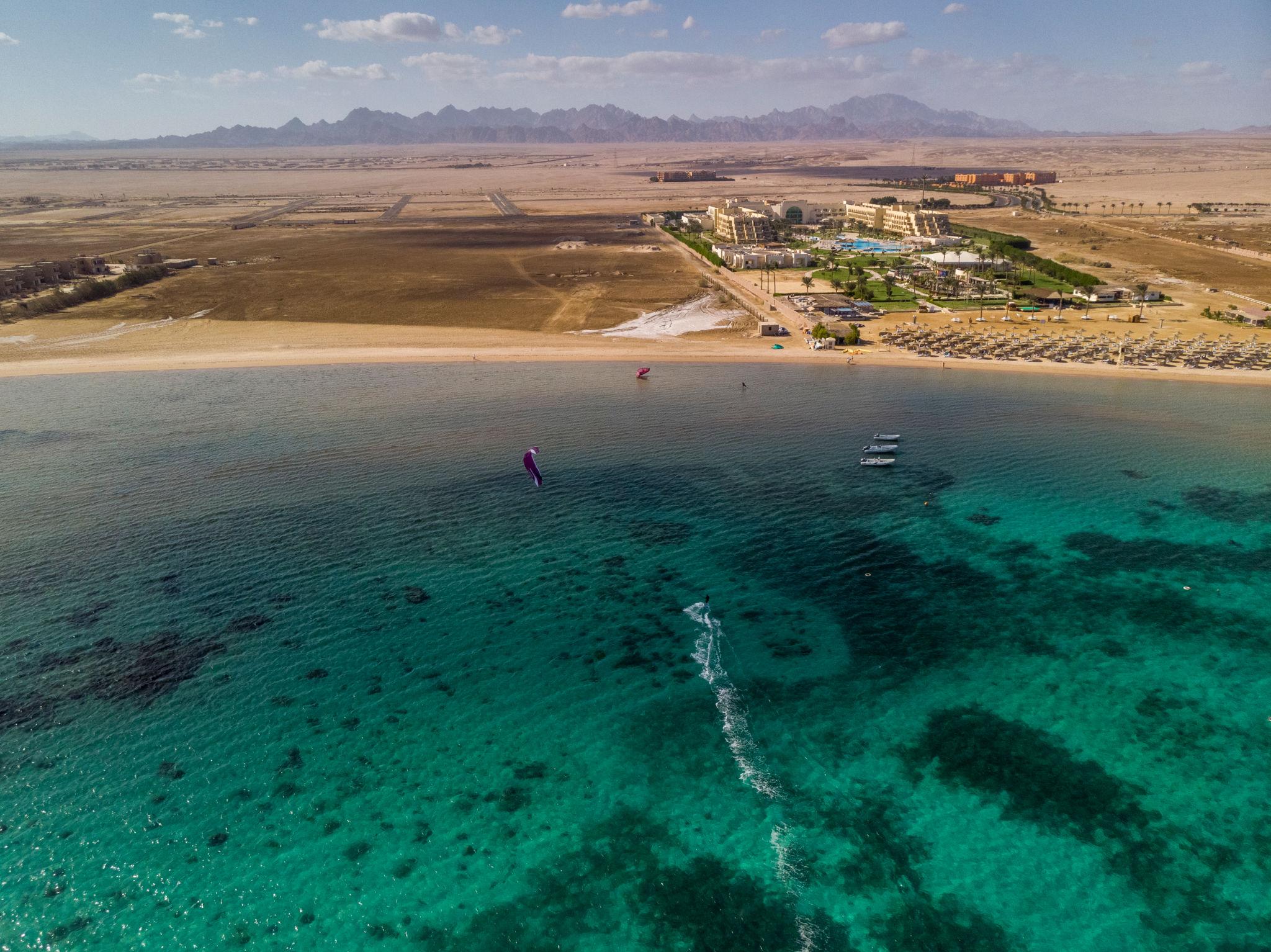 Aerial view of a kitesurfer on vivid turquoise Red Sea waters beside a desert resort beach.