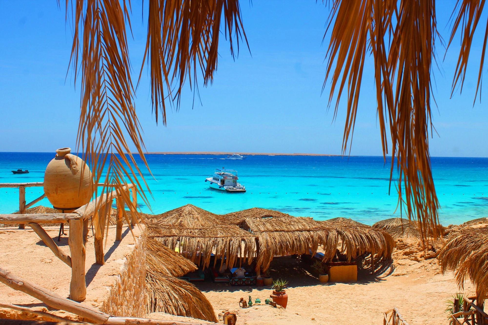 Thatched beach huts on golden sand overlooking vivid turquoise Red Sea waters with a boat anchored offshore