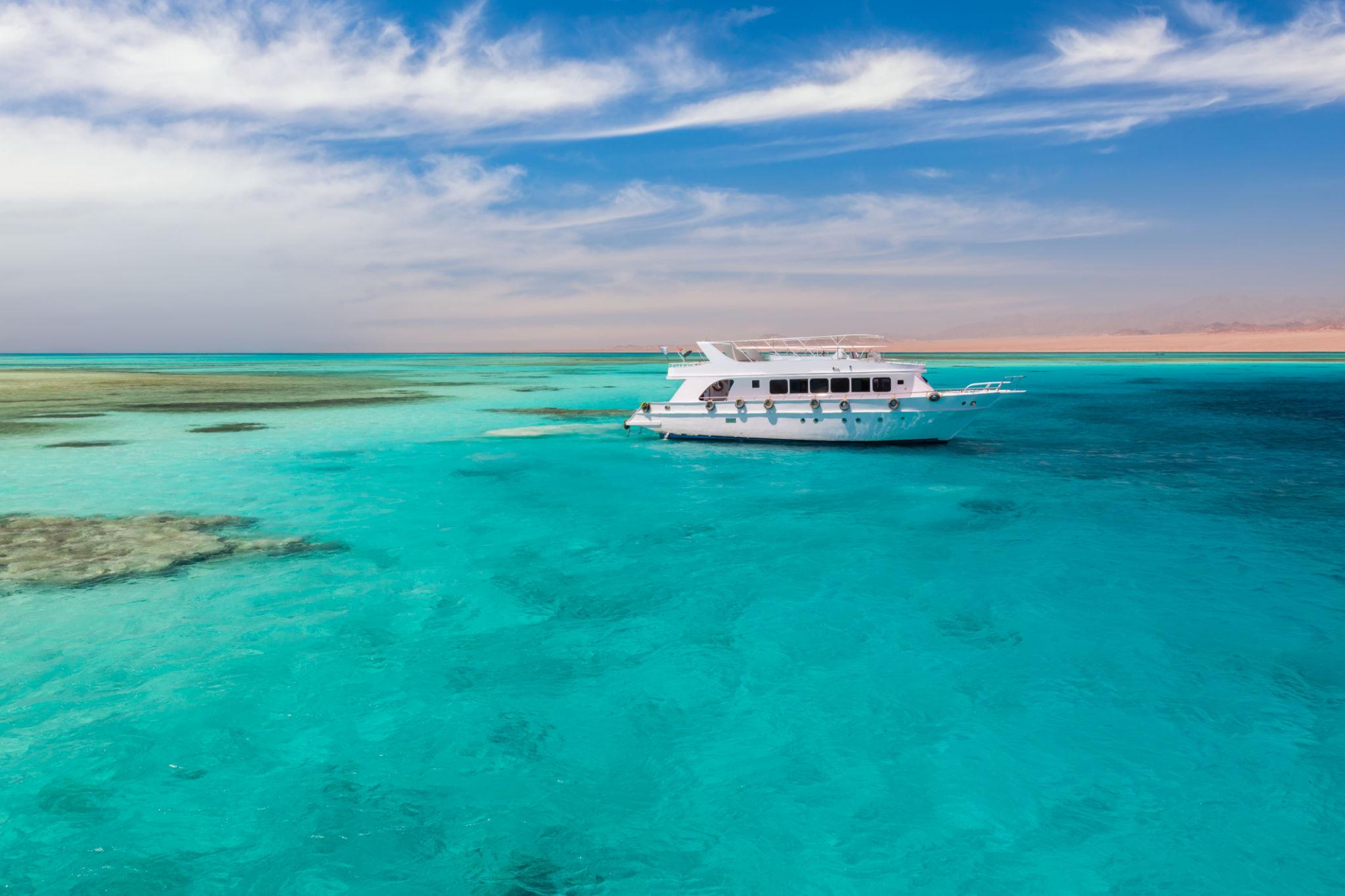A white yacht anchored over a vivid turquoise coral reef with arid hills beyond
