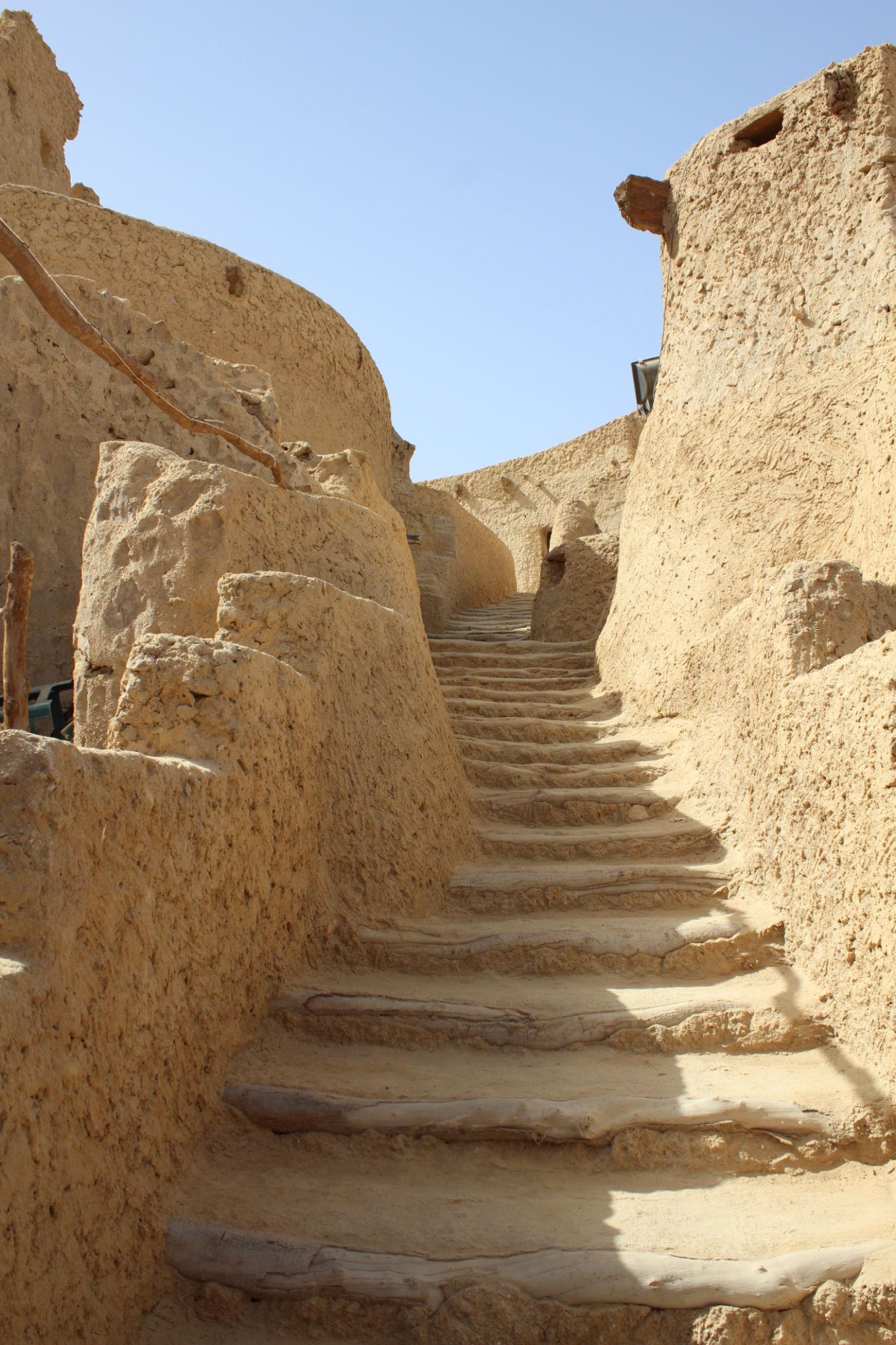 Narrow stone steps rising between eroded mud-brick walls in Siwa's old town under a clear blue sky