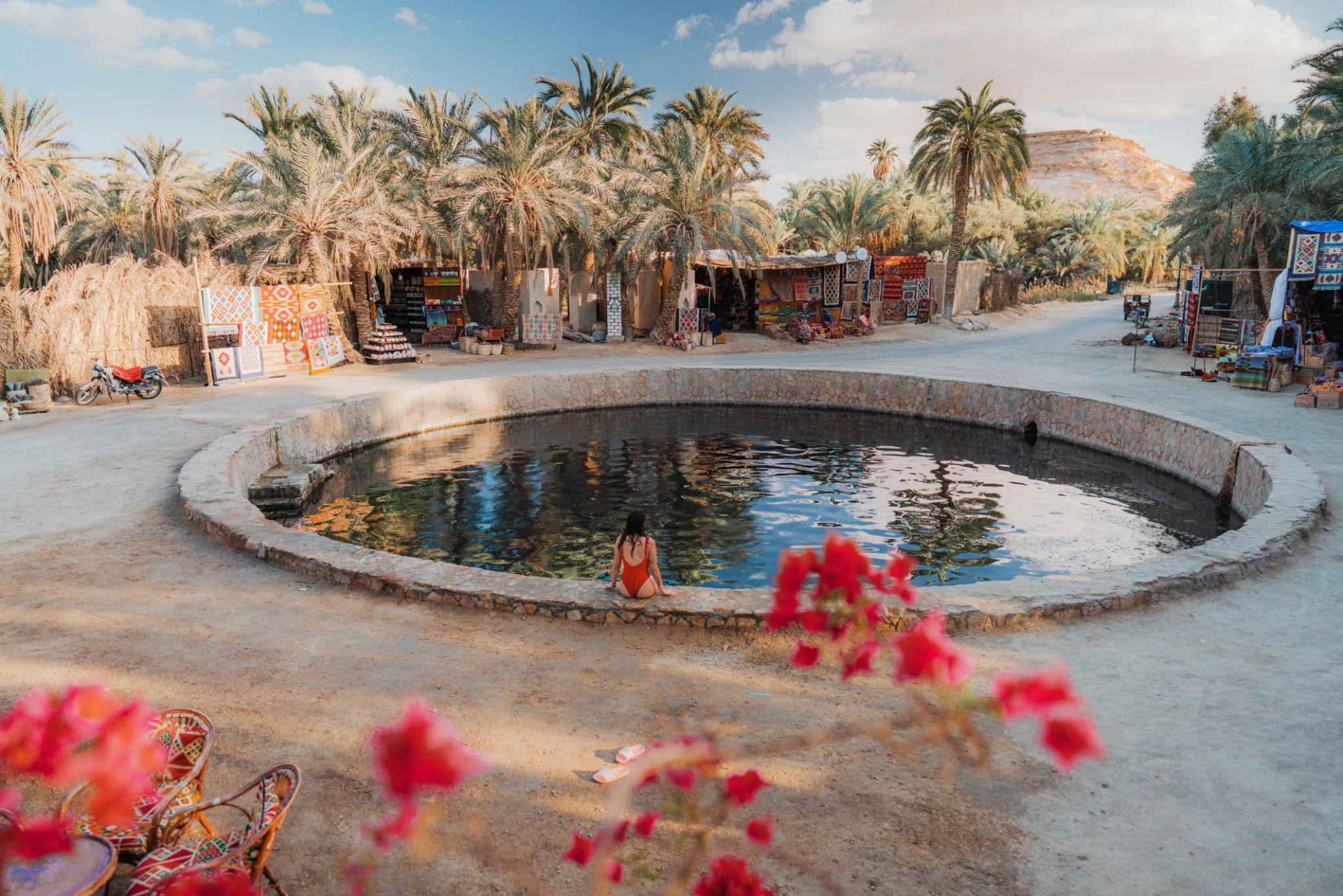 A woman in a red swimsuit sits at the edge of a circular natural spring pool surrounded by palm trees and market stalls