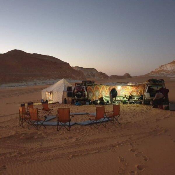 A glowing desert camp with circle of chairs and two 4x4 vehicles at dusk in Siwa.