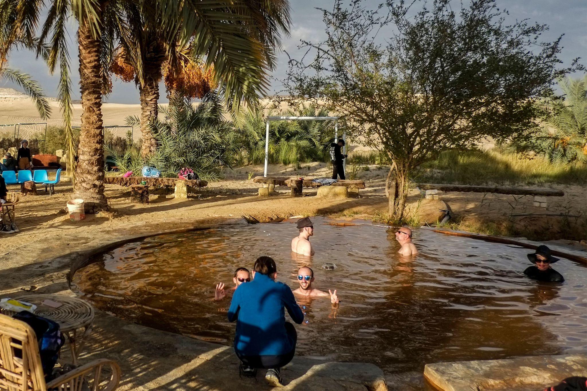 Tourists bathing in a natural hot spring pool surrounded by palm trees and sand dunes