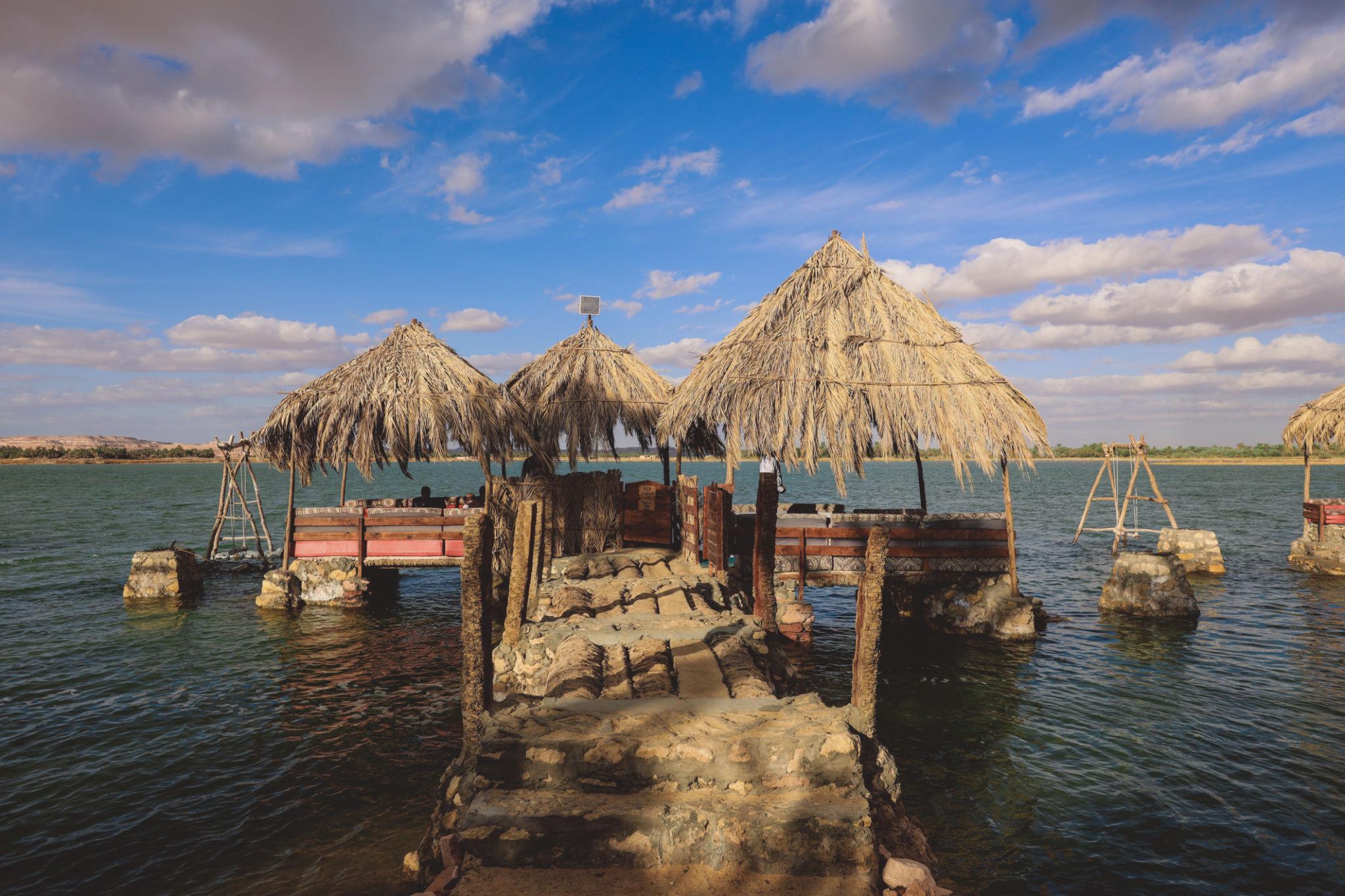 Rustic thatched-roof huts on a stone platform over calm lake waters in Siwa Oasis