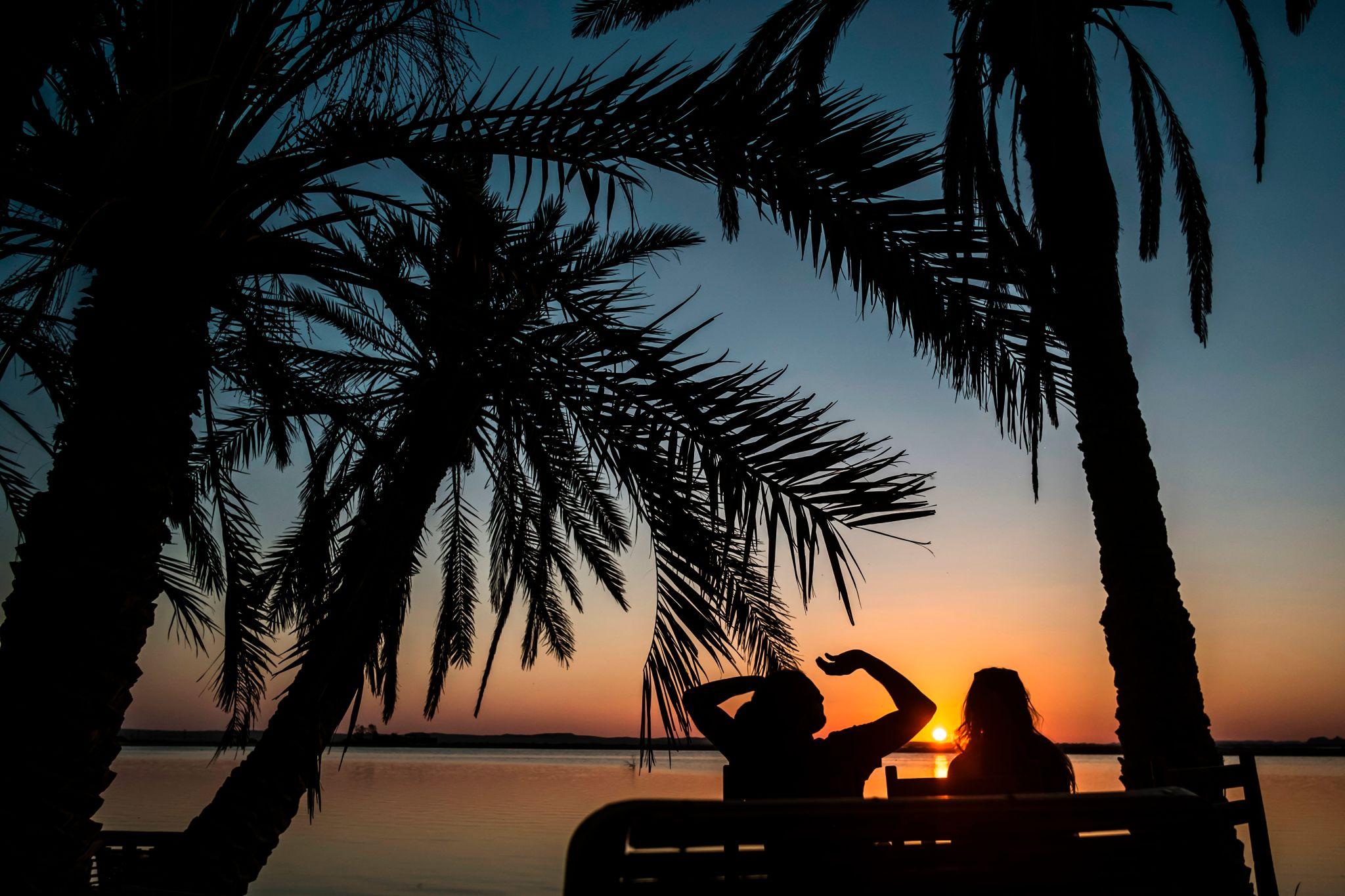 Two people silhouetted against a vivid sunset over Siwa Oasis lake, framed by palm trees