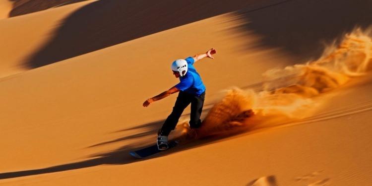 A helmeted sandboarder carves down a steep golden sand dune kicking up a spray of sand