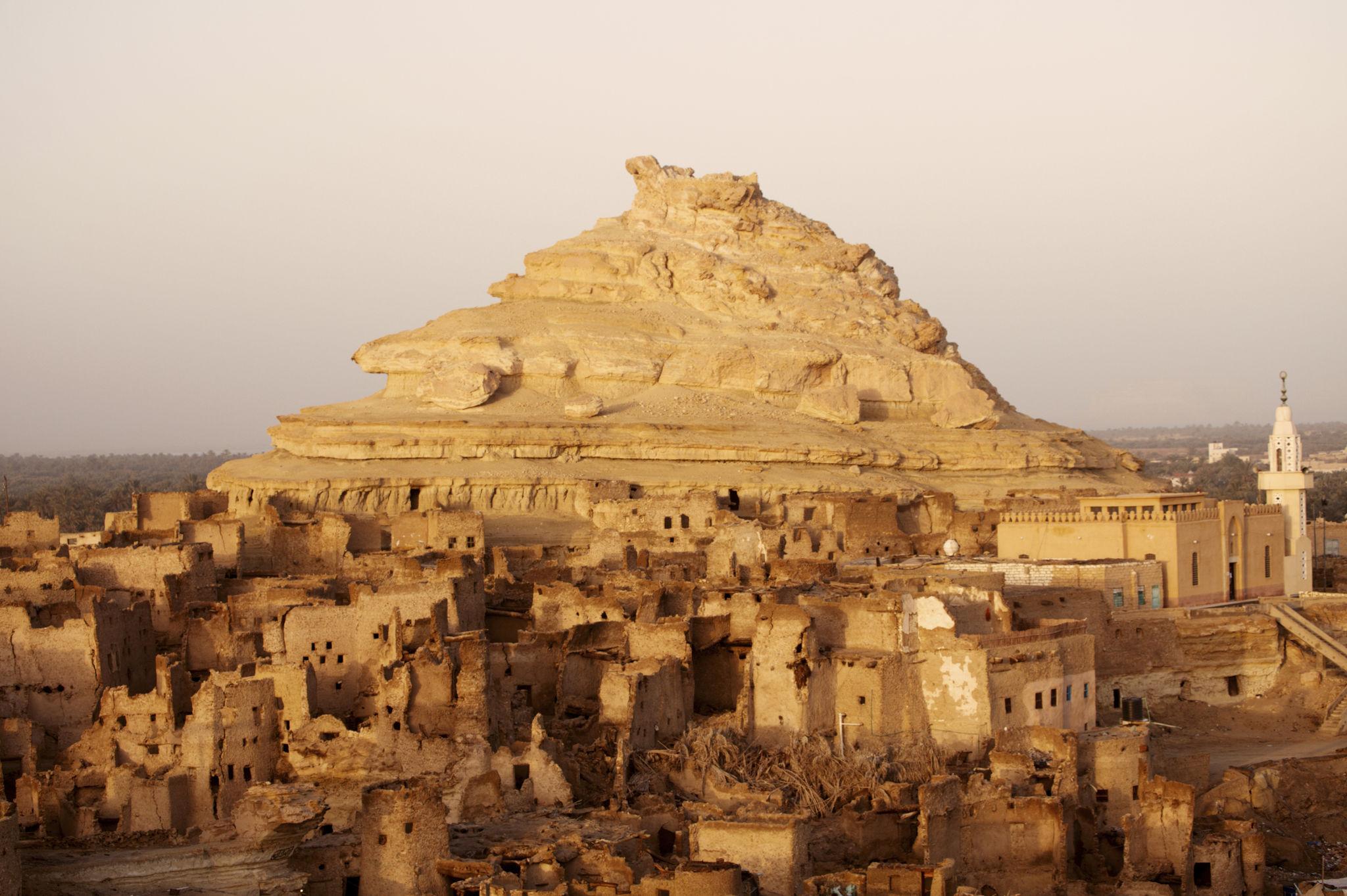 Ancient mud-brick ruins of Shali fortress beneath a layered sandstone rock formation in Siwa Oasis.