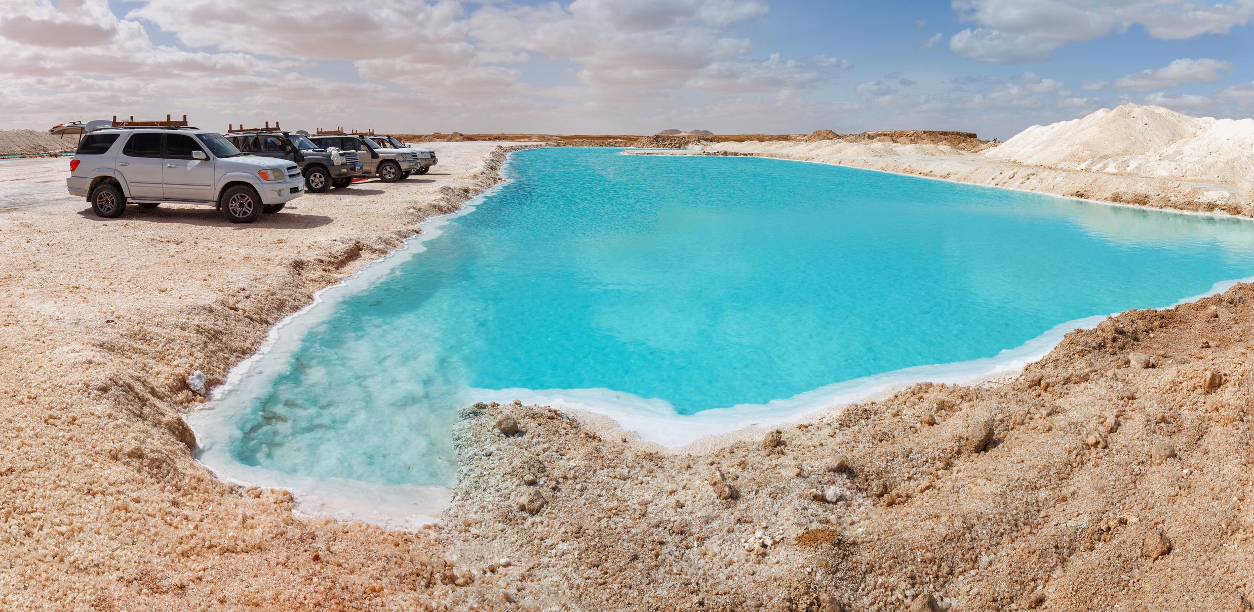 Vivid turquoise salt lake surrounded by white salt flats and parked 4x4s in Siwa Oasis