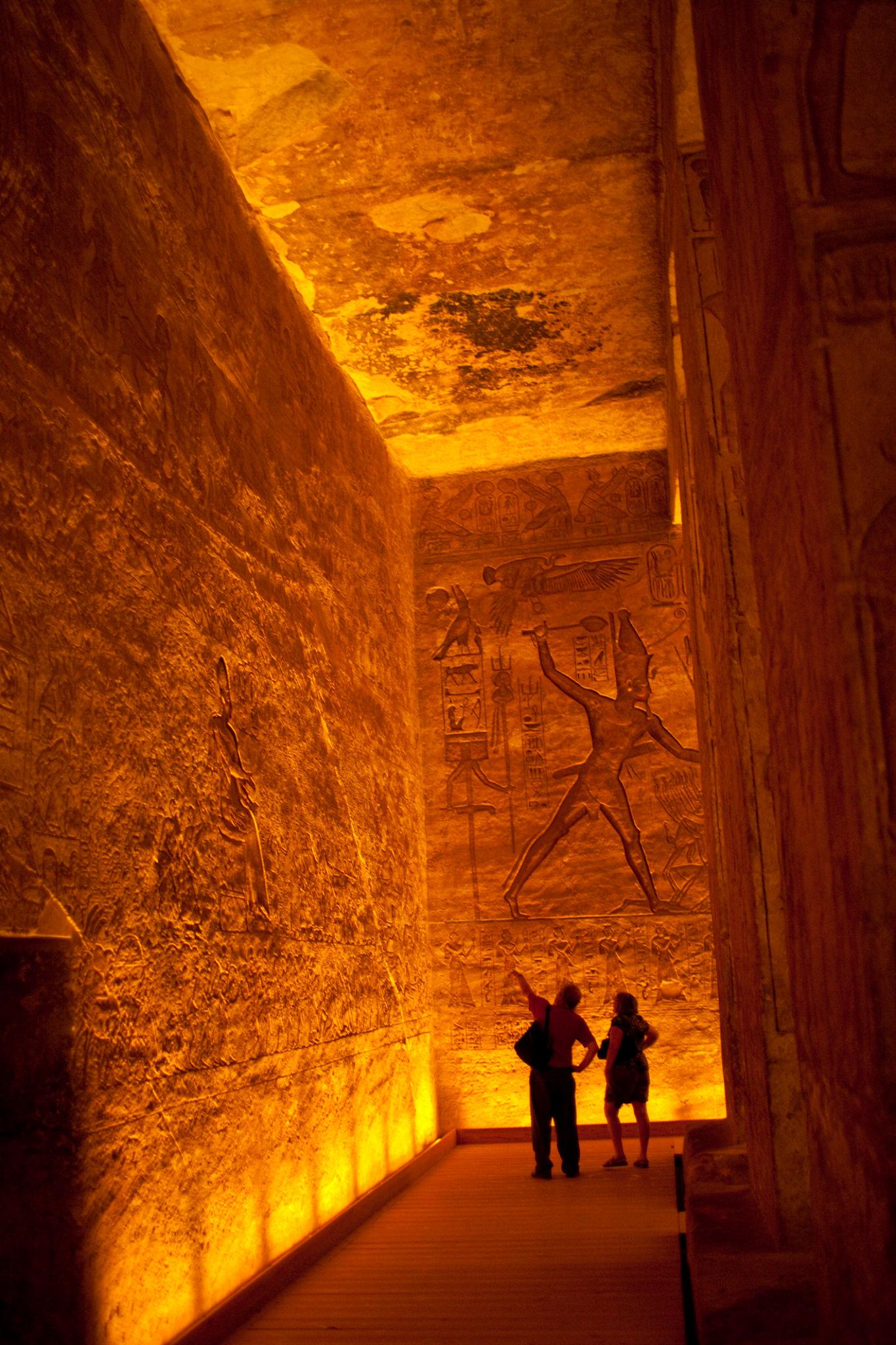 Two tourists silhouetted against golden-lit ancient Egyptian relief carvings inside Abu Simbel temple