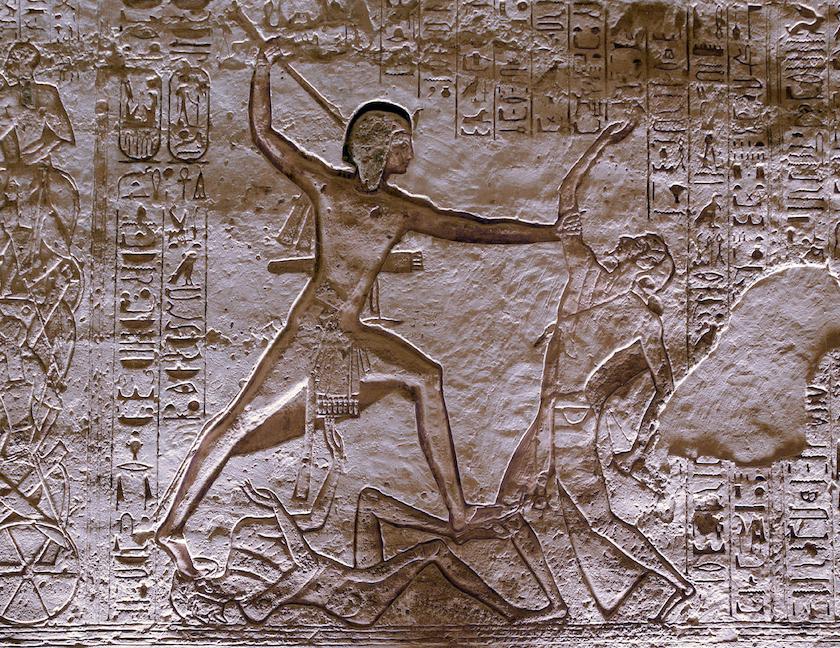 Ancient Egyptian bas-relief carving of a pharaoh striking enemies, surrounded by hieroglyphs.