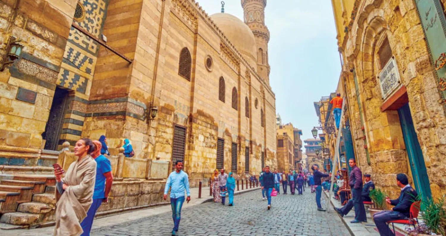 Pedestrians walk along a cobblestone street lined with grand Islamic stone monuments in Cairo