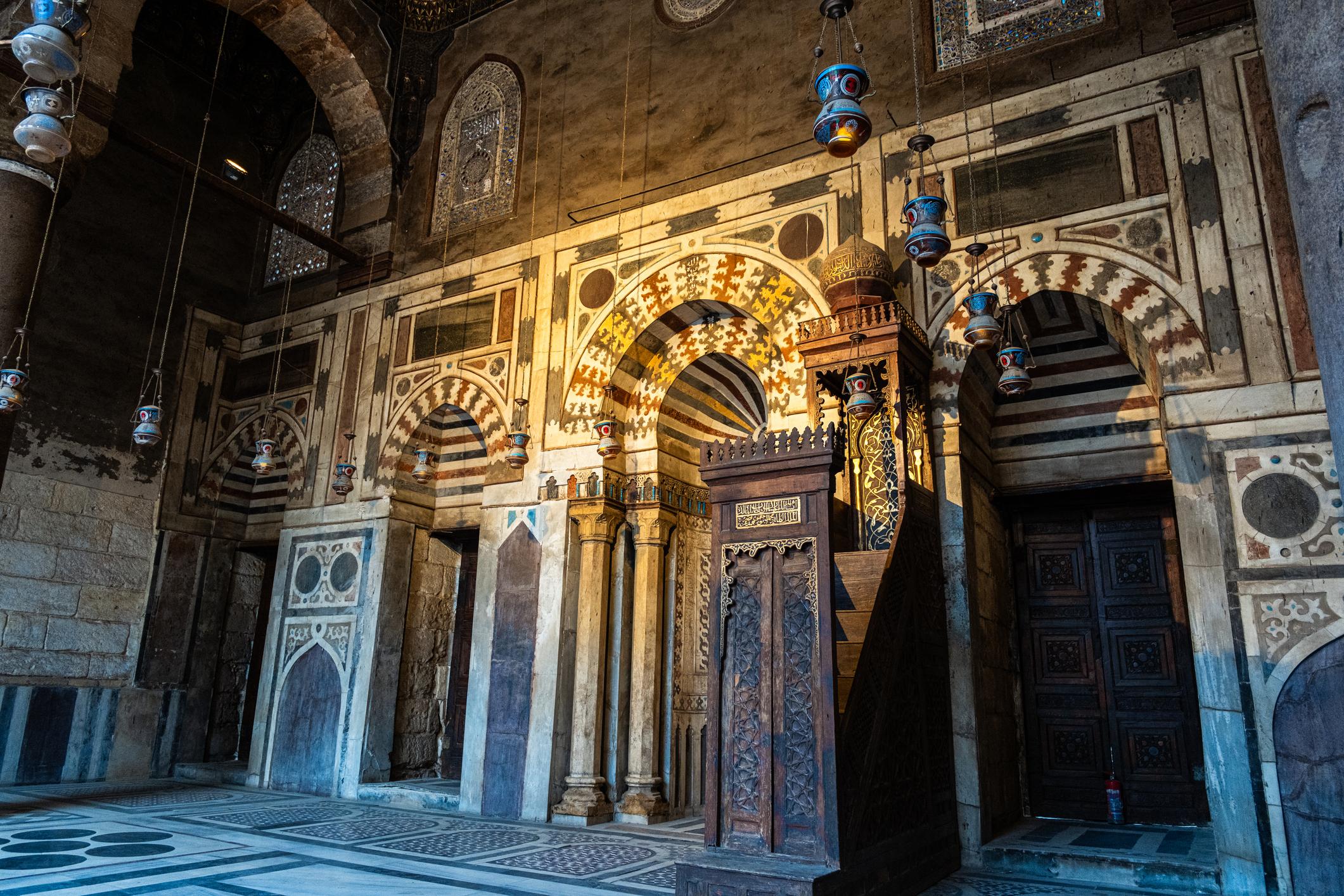 Ornate mihrab and carved wooden minbar inside a historic Cairo mosque with hanging lanterns