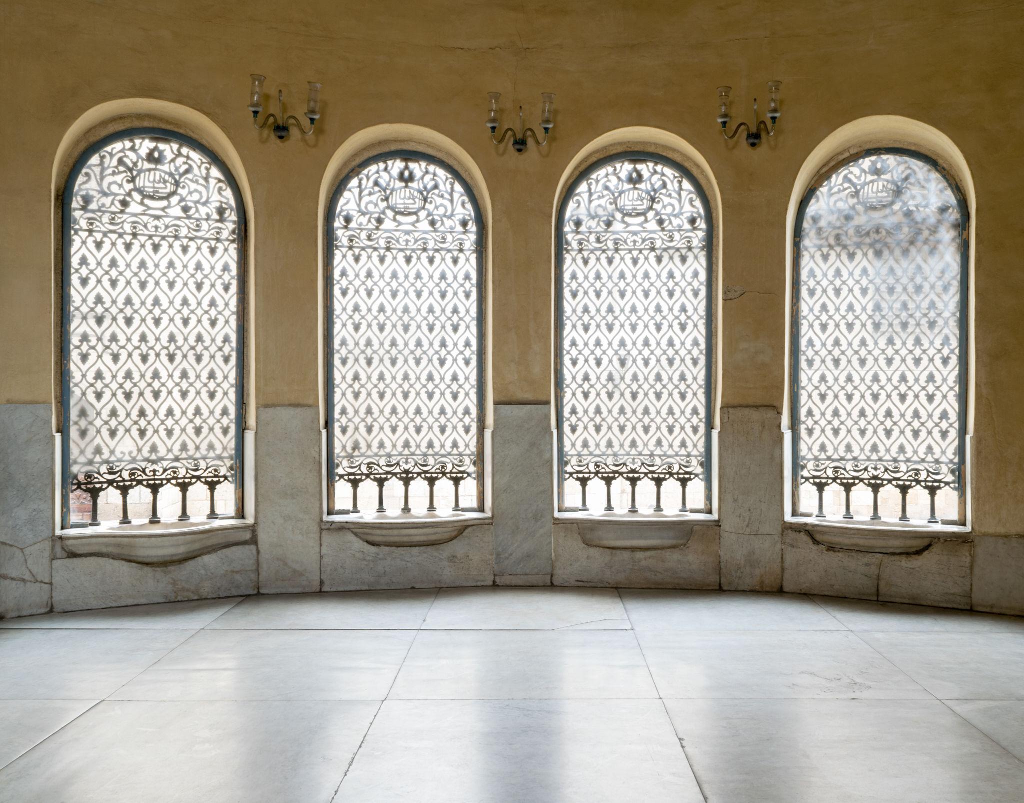 Four tall arched windows with decorative iron lattice grilles in a marble-floored hall at NMEC