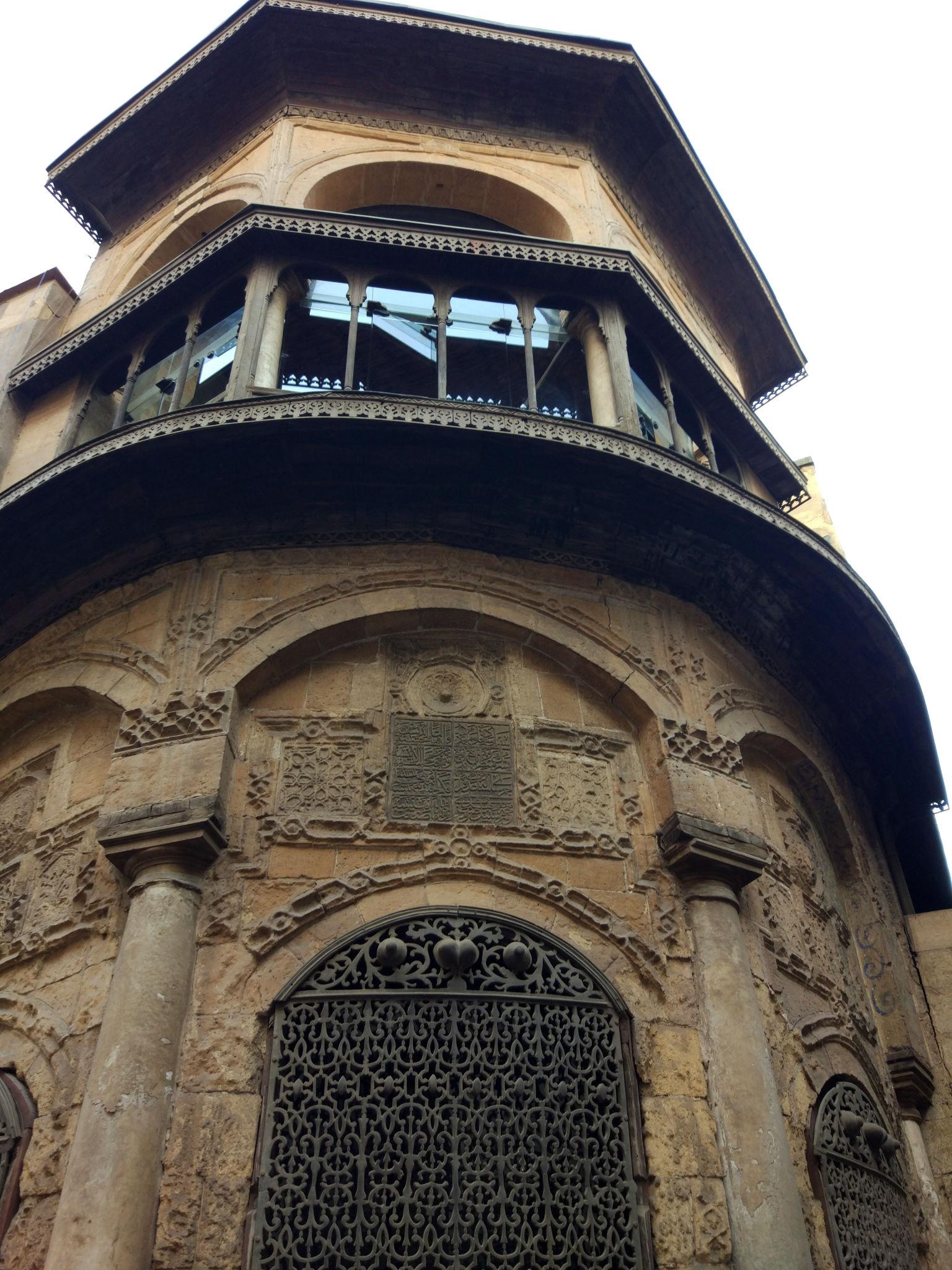Looking up at an ornate Islamic stone tower with carved balcony and decorative iron gate entrance