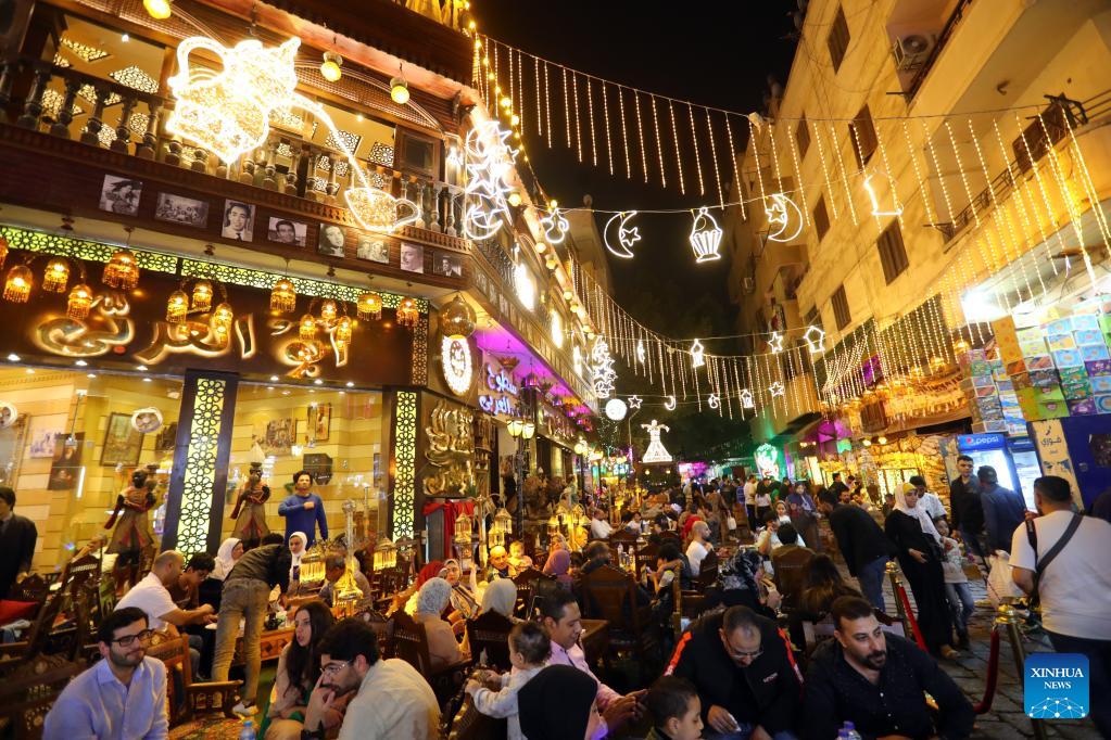 Crowds gather beneath glowing Ramadan lights and crescent decorations on a busy Cairo street at night.