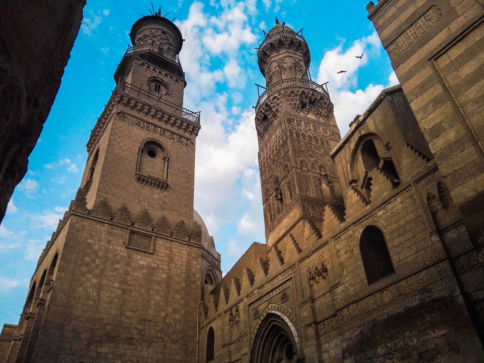 Two ornate Mamluk minarets rising above an intricately carved stone facade against a blue sky