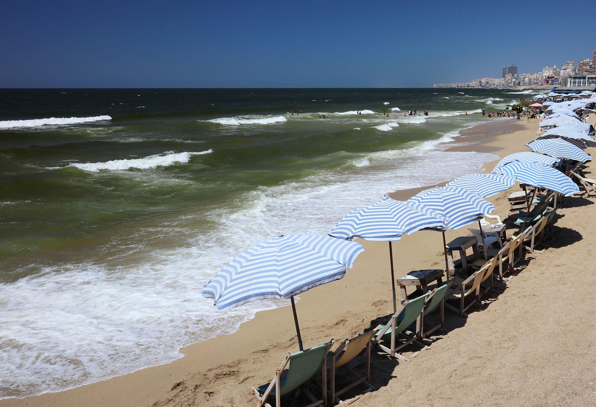 Row of blue-striped beach umbrellas and loungers on a sandy Corniche beach along the Mediterranean