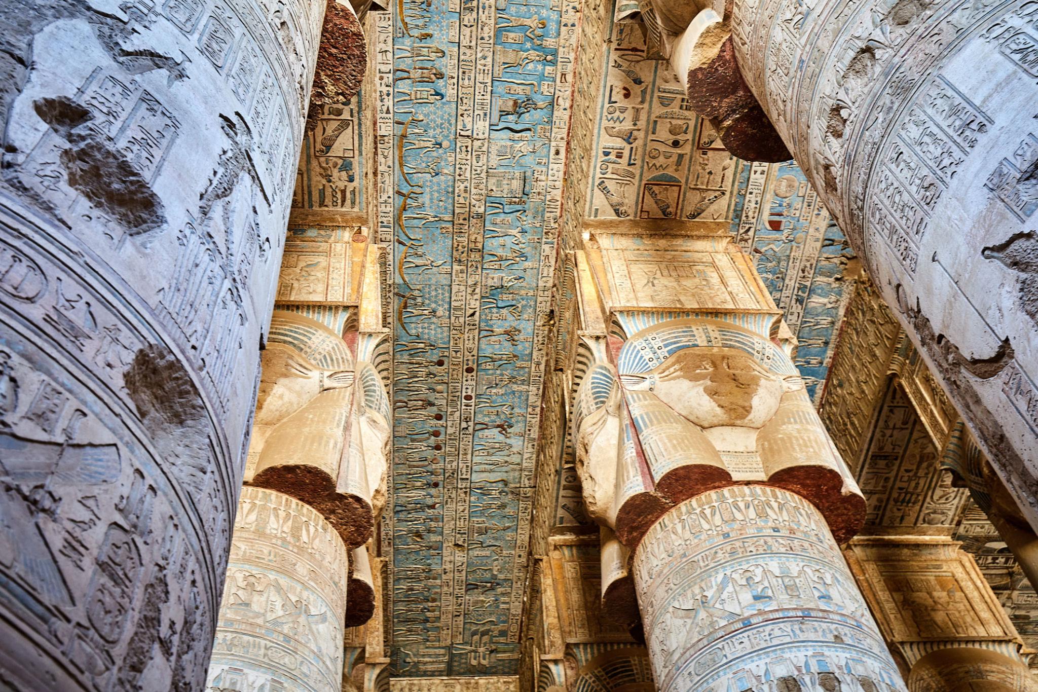 Upward view of Hathor capitals and painted ceiling beams inside Dendera Temple.