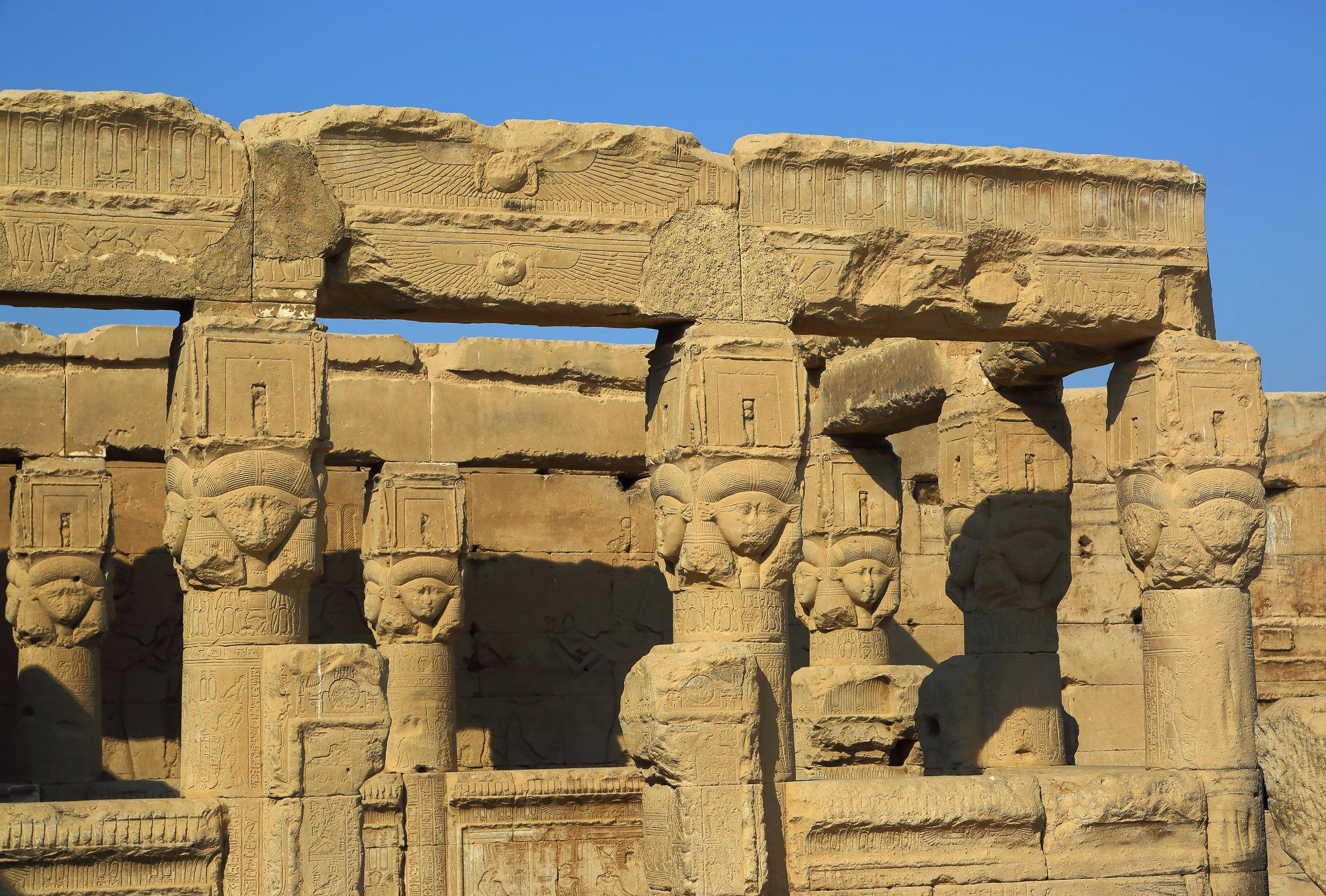 Exterior row of sandstone Hathor-headed capitals against a clear blue sky.
