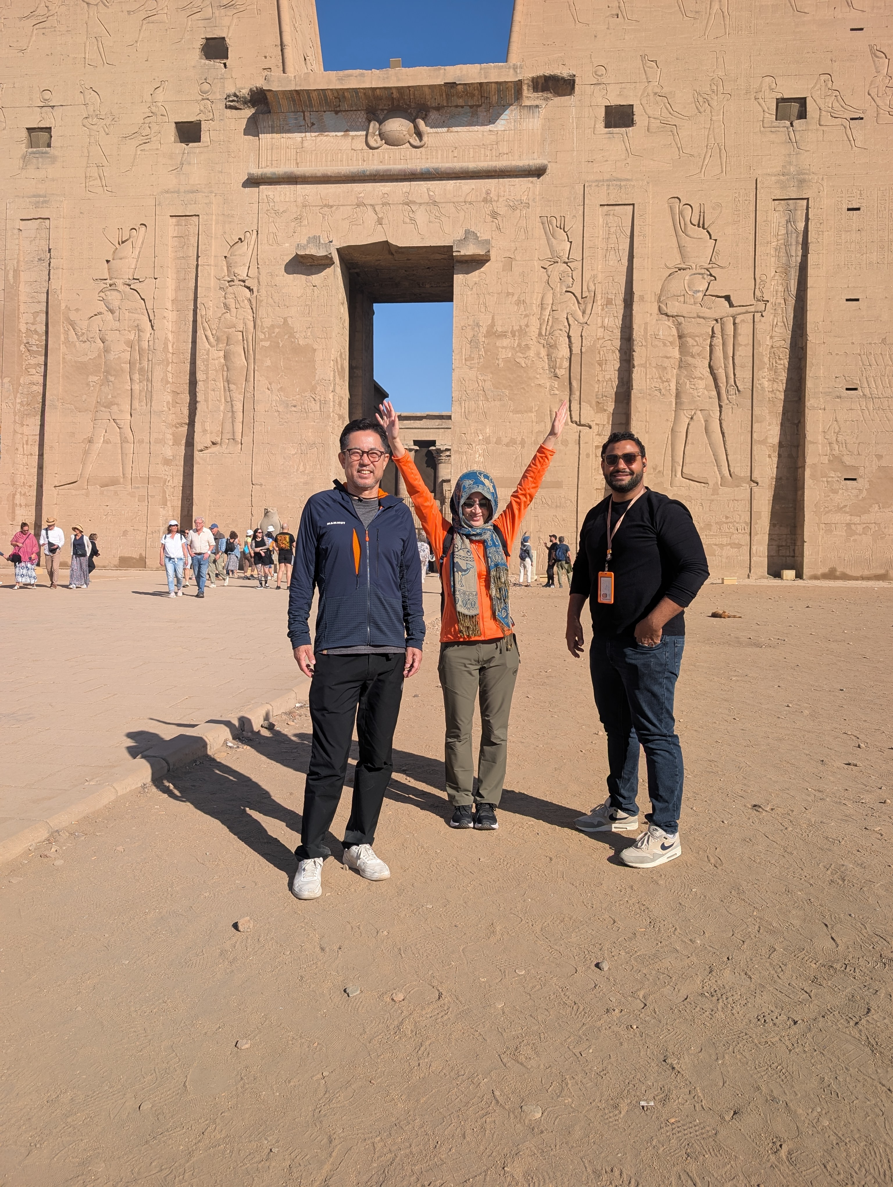 Three travellers celebrating with raised arms before the Temple of Edfu pylon.