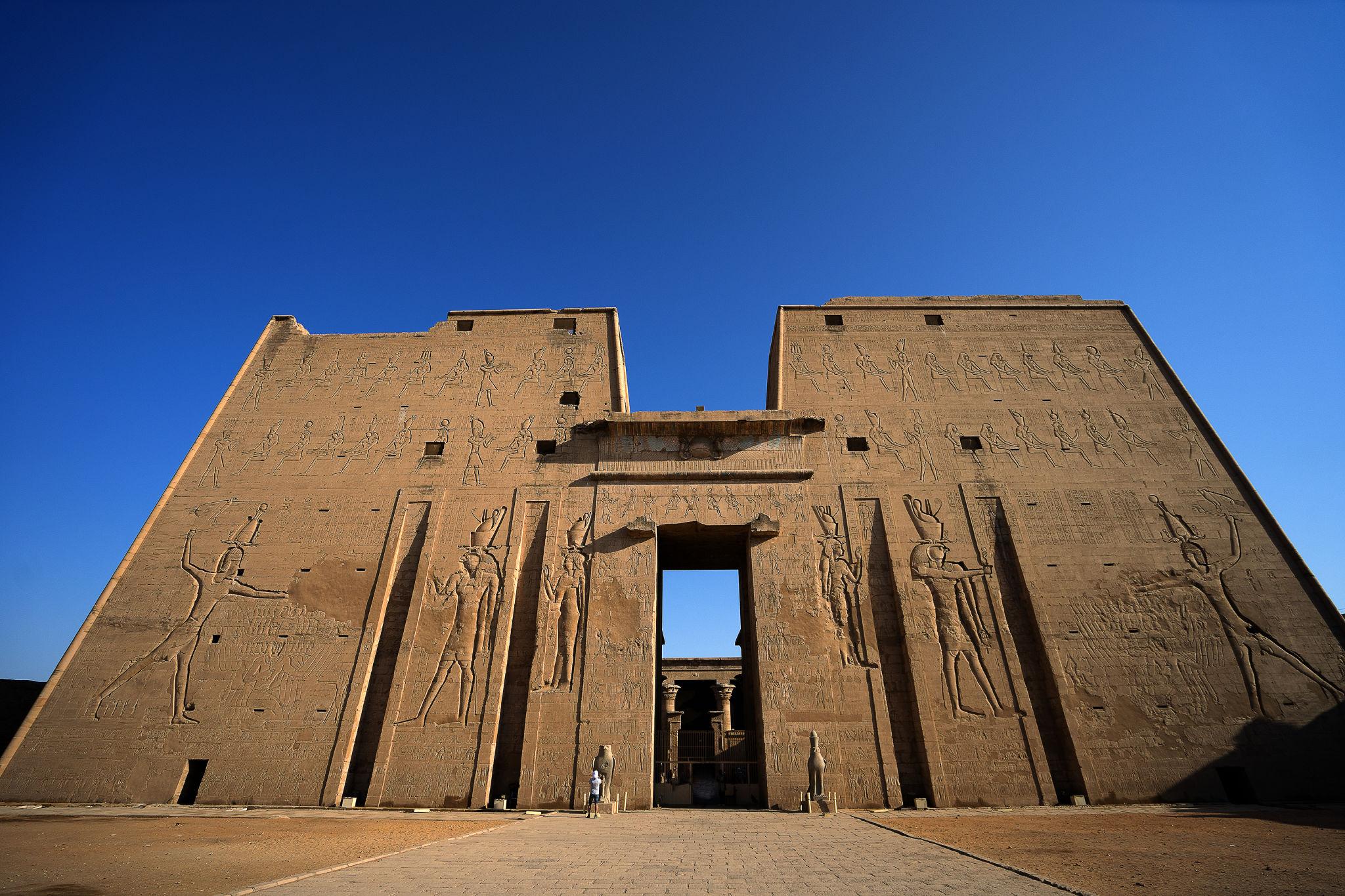Wide view of the Temple of Edfu pylon entrance with tiny visitors for scale.