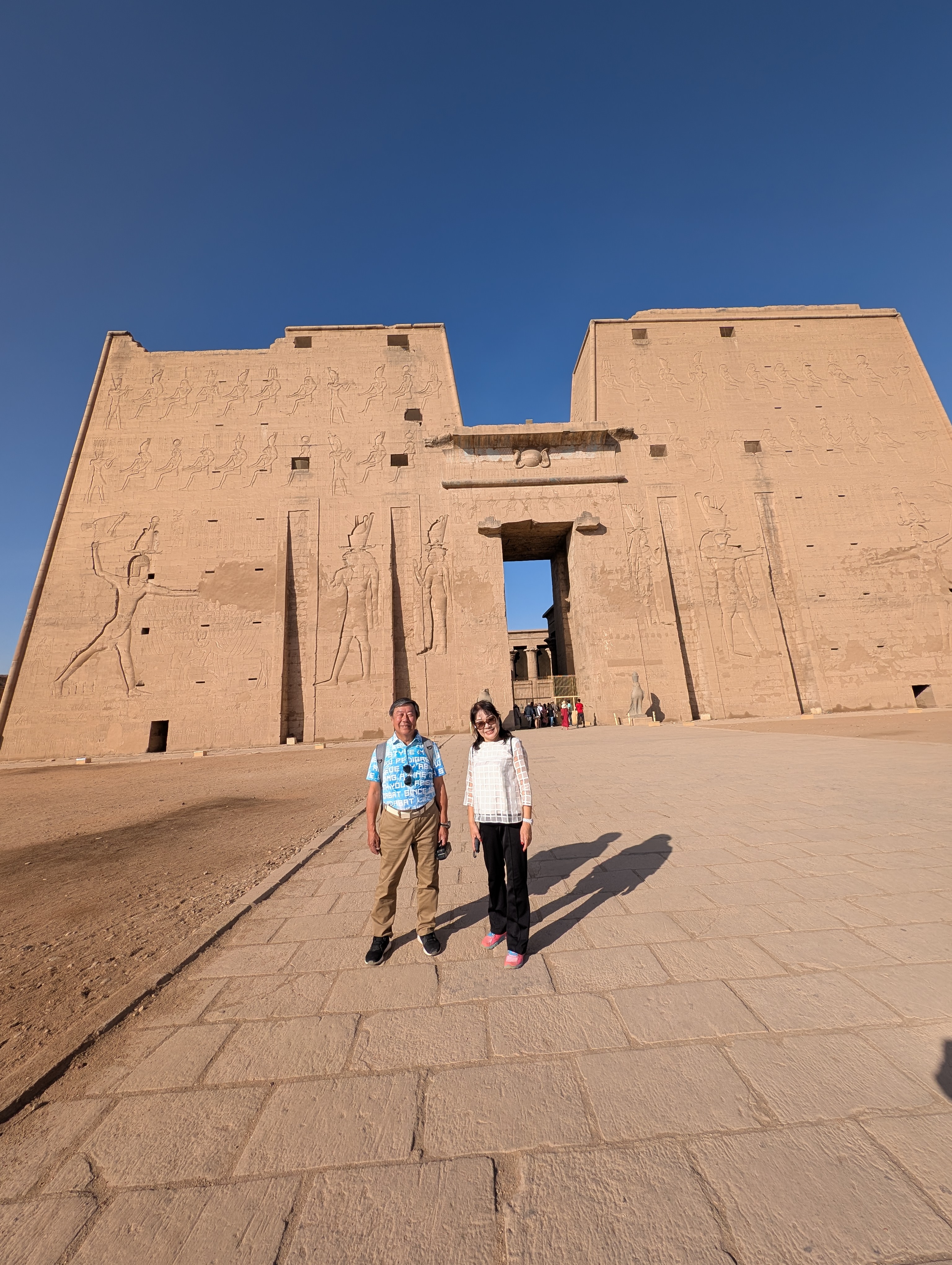 Two visitors standing on the paved plaza before the Temple of Edfu pylon.